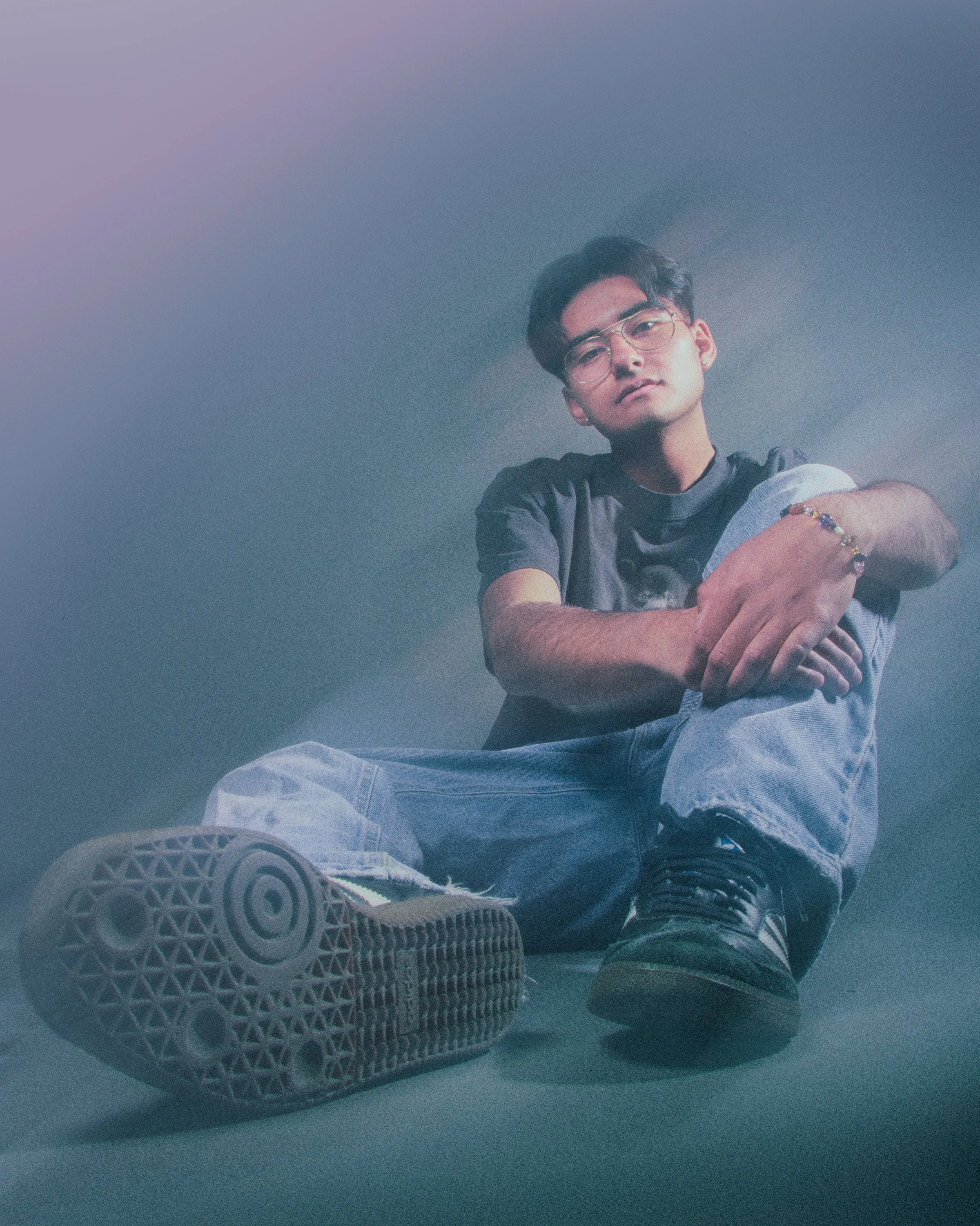 Young man sitting on the floor with legs stretched out, wearing glasses, a black T-shirt, ripped jeans, and black sneakers, looking at the camera with a neutral expression, surrounded by a soft, muted lighting.