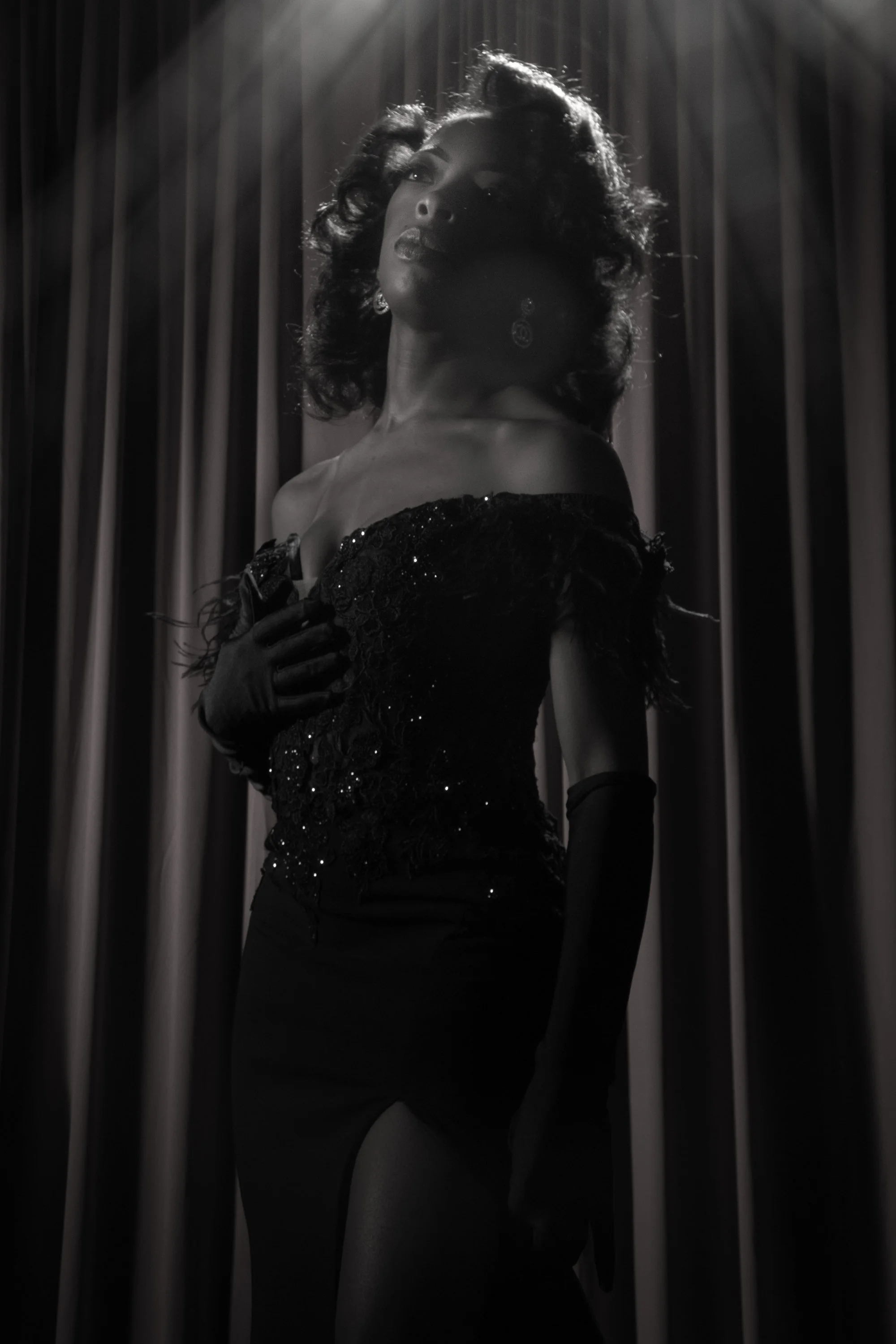 Black and white photo of a woman with curly hair, wearing a vintage-style dress with sequins, gloves, and earrings, standing in front of curtains with lighting highlighting her face and upper body.
