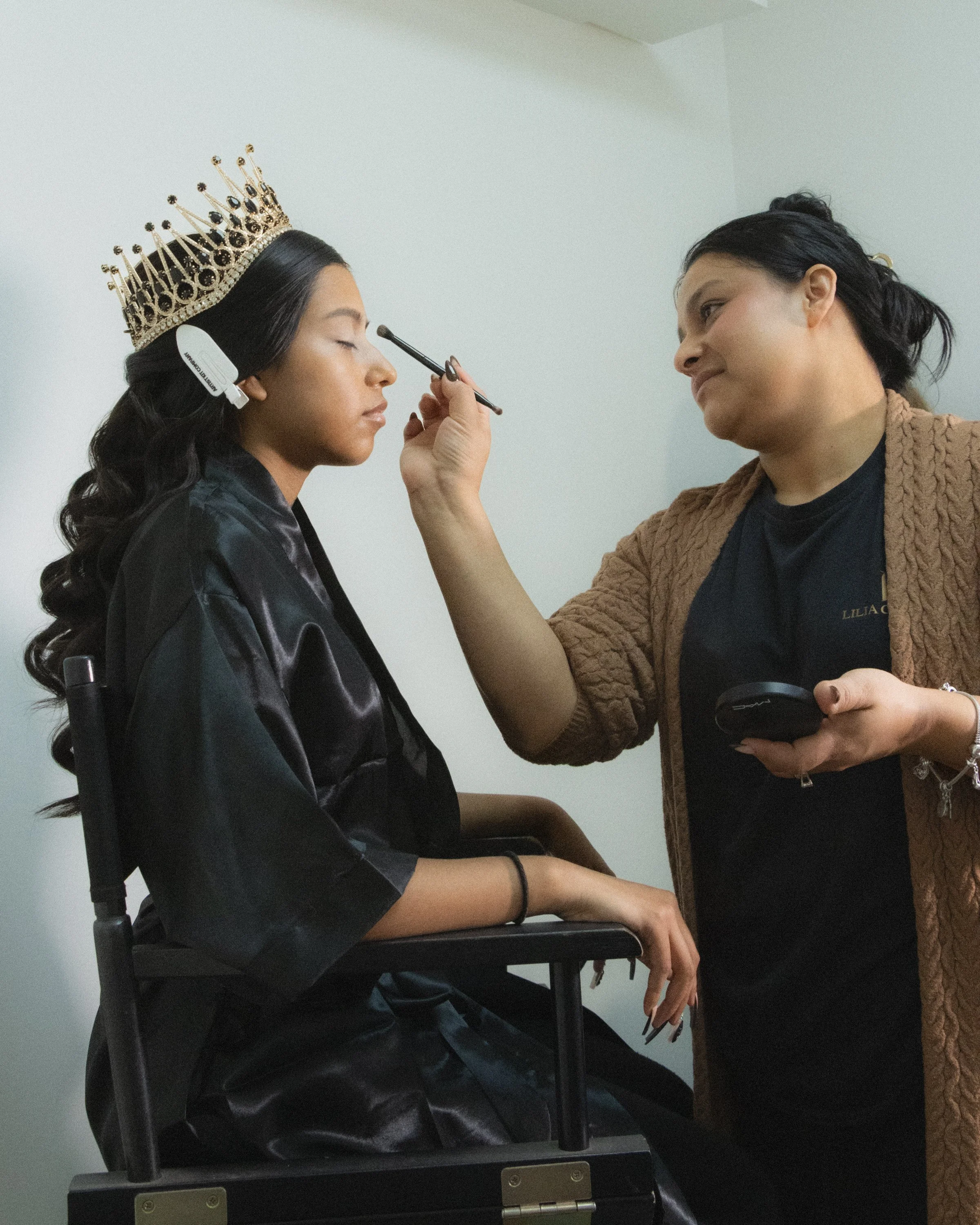 A woman sitting with eyes closed while another woman applies makeup with a brush, wearing a crown and a hair clip.