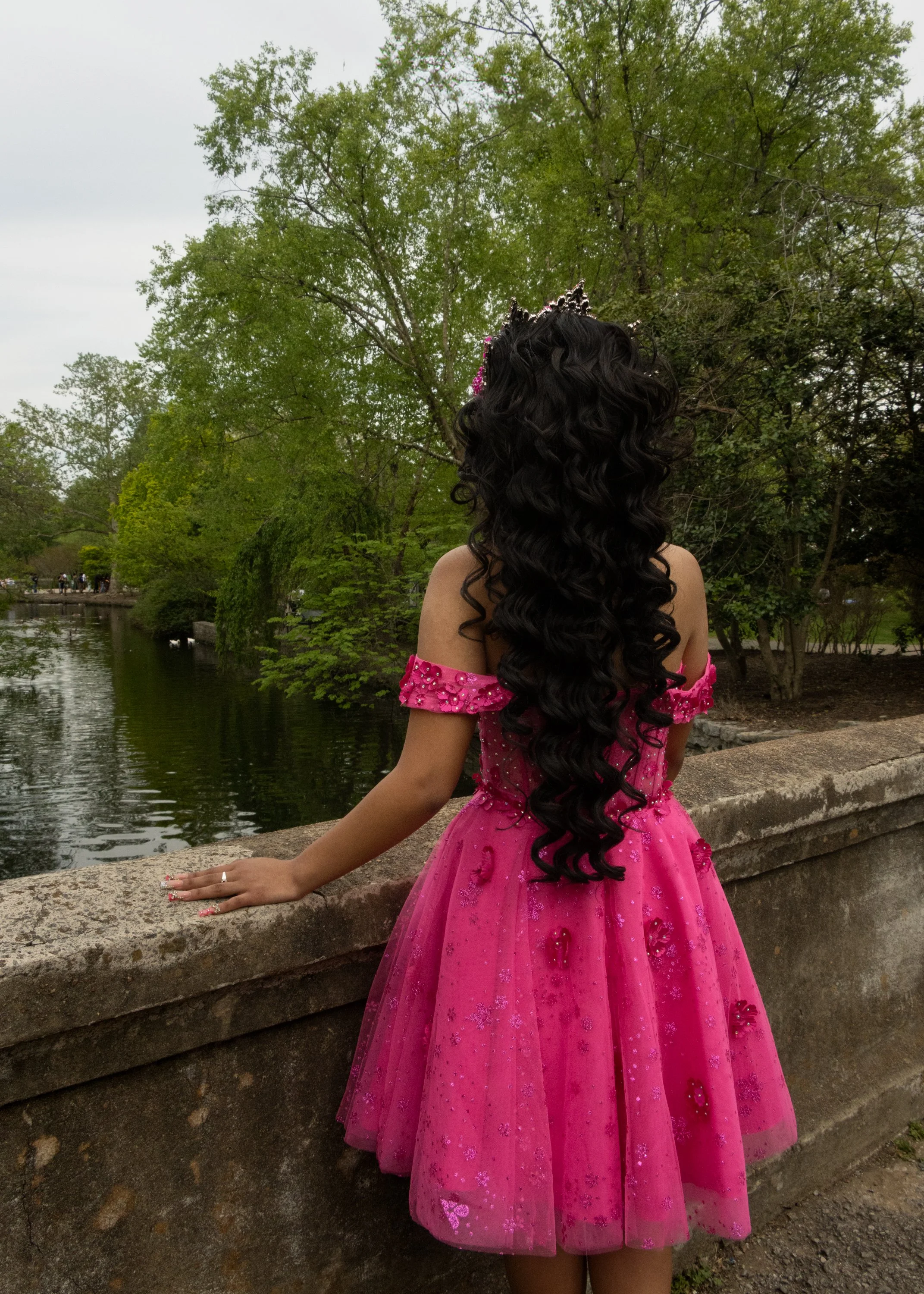 A woman in a bright pink dress with embellishments, long curly black hair, and a small crown on her head, standing by a stone ledge near a river in a park with green trees.