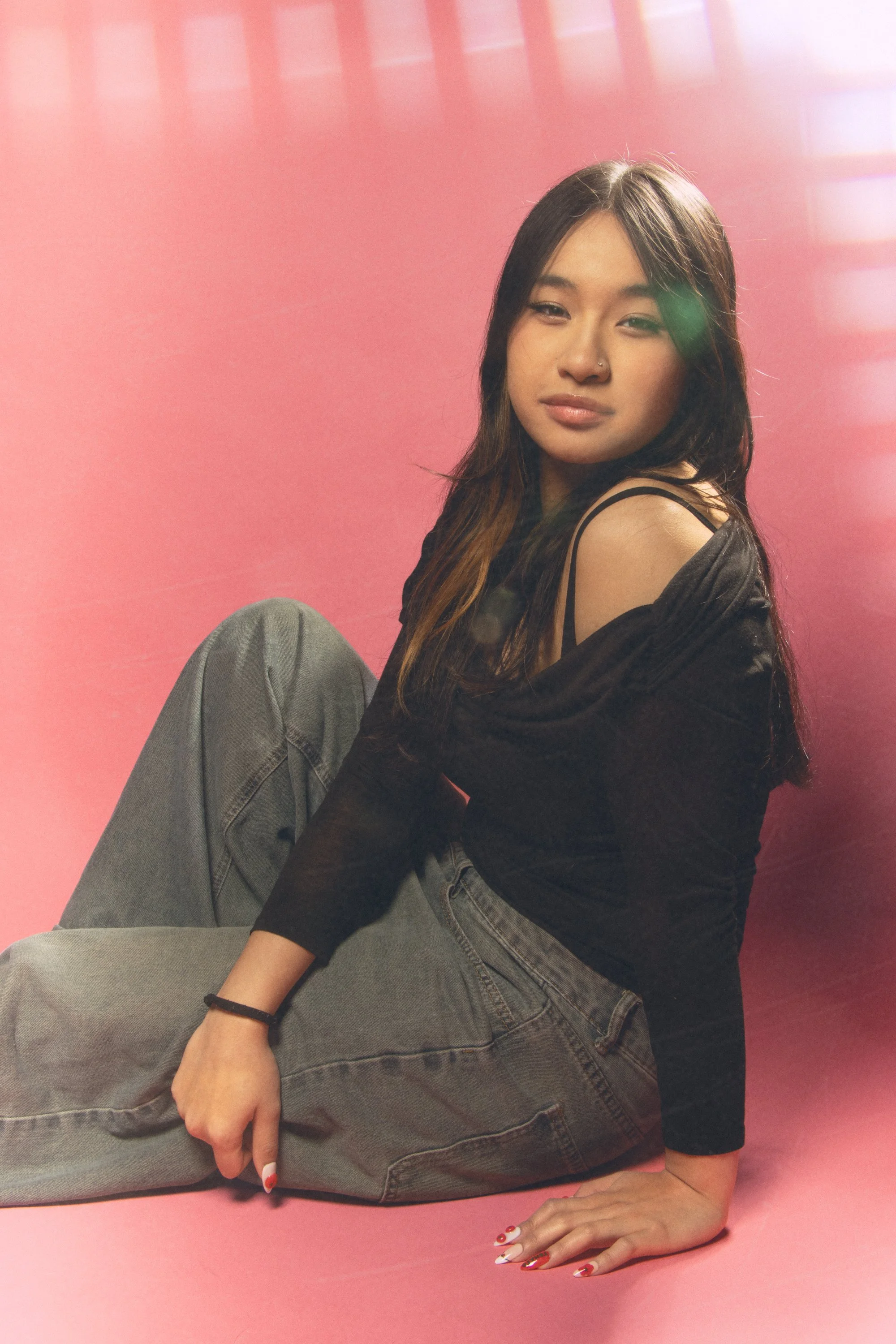 A young woman with long dark hair sitting on the floor against a pink backdrop, wearing a black top and gray jeans, with one arm resting on her knee and the other on the floor, displaying manicured nails with a holiday design.