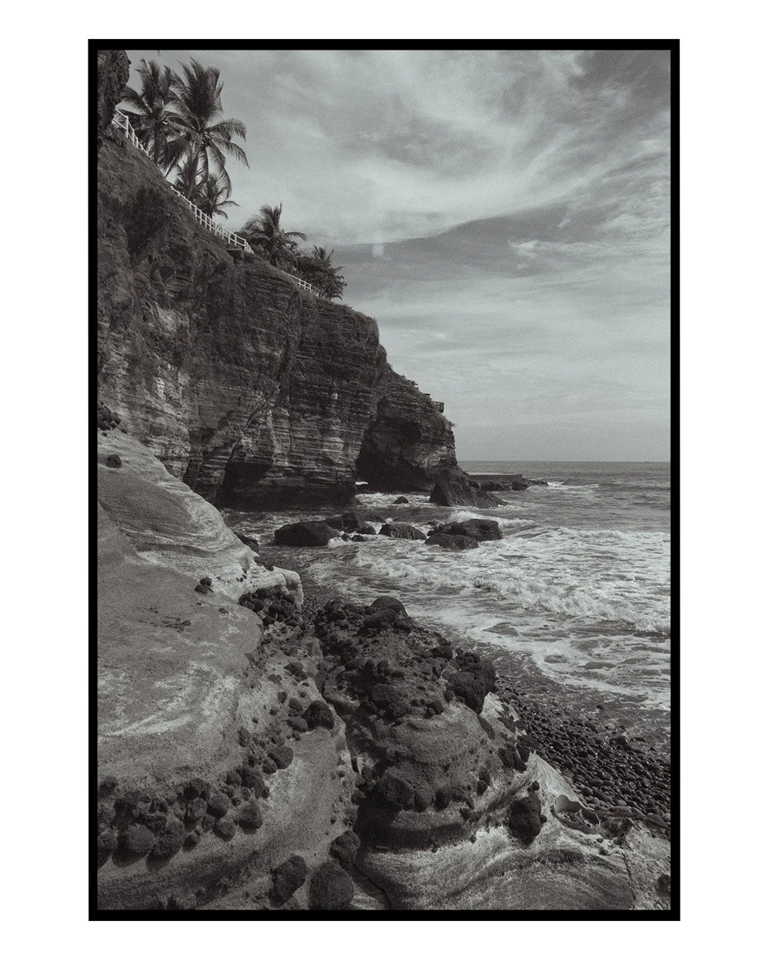 Black and white photograph of a rocky coastal area with cliffs, palm trees at the top of the cliffs, and waves crashing against the rocks below.