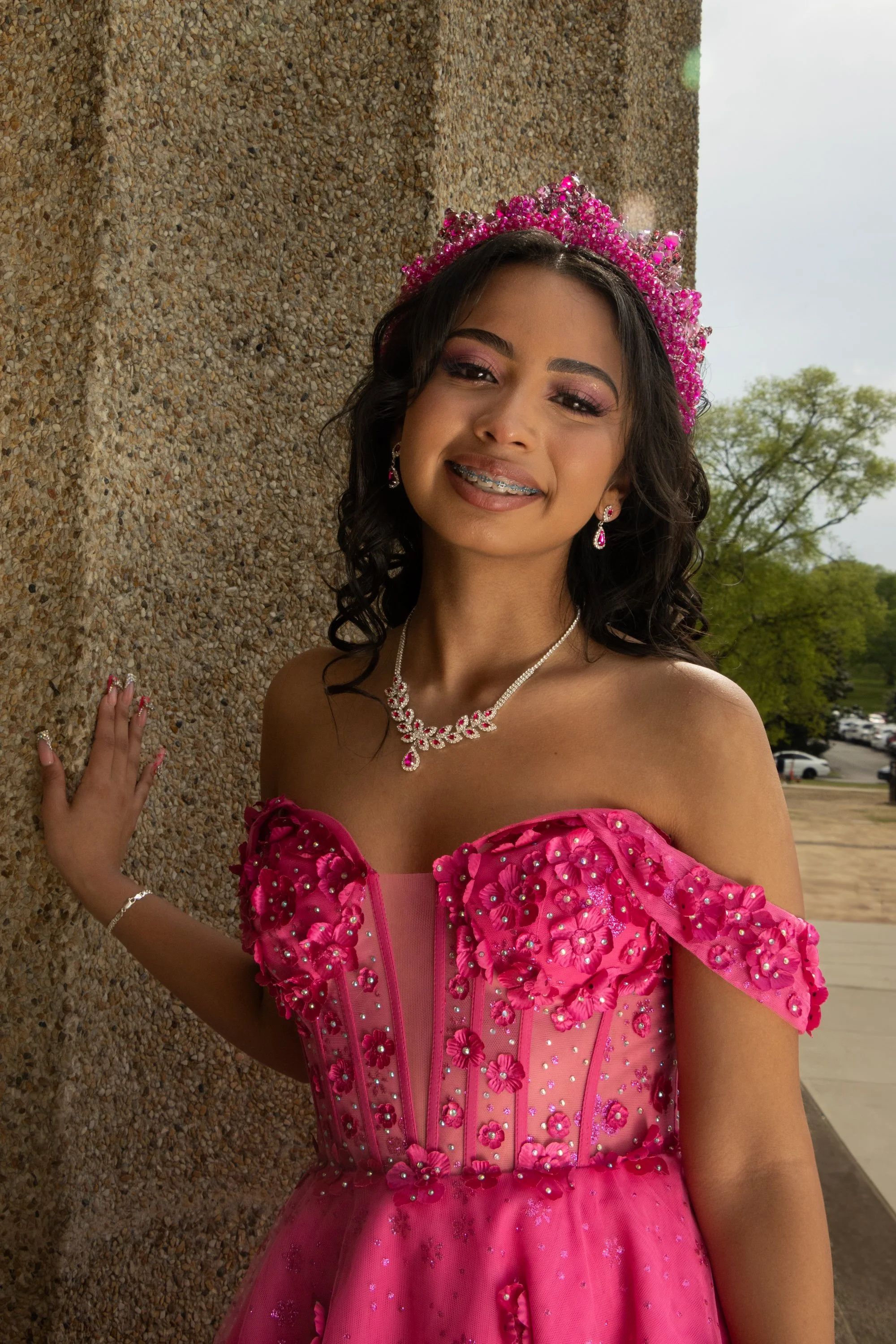 A young woman dressed in a pink gown with floral embellishments and matching jewelry, wearing a pink tiara, standing outdoors near a textured wall, with trees and parked cars visible in the background.