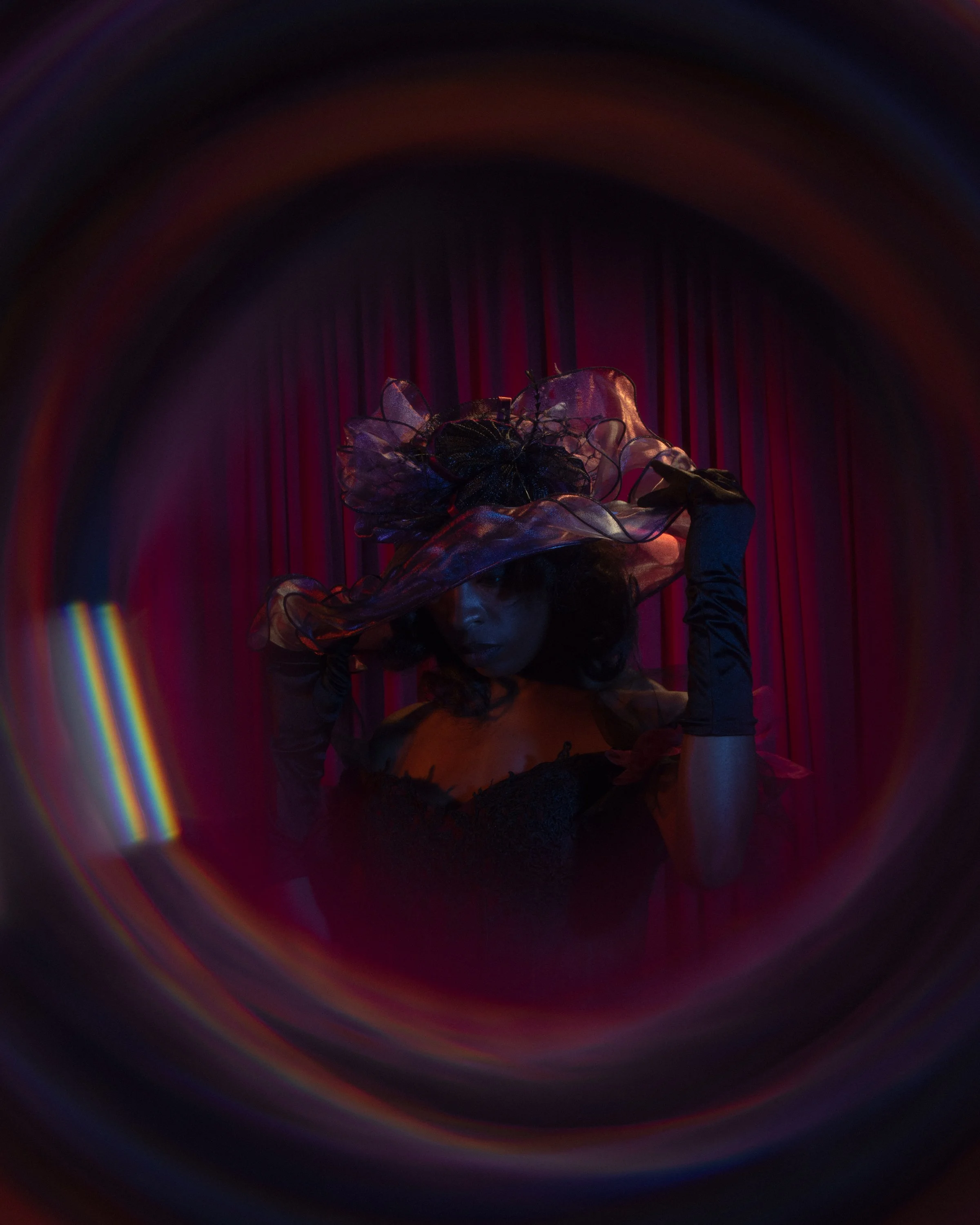 A woman on stage wearing an elaborate, artistic headpiece and black gloves, with a red velvet curtain in the background.