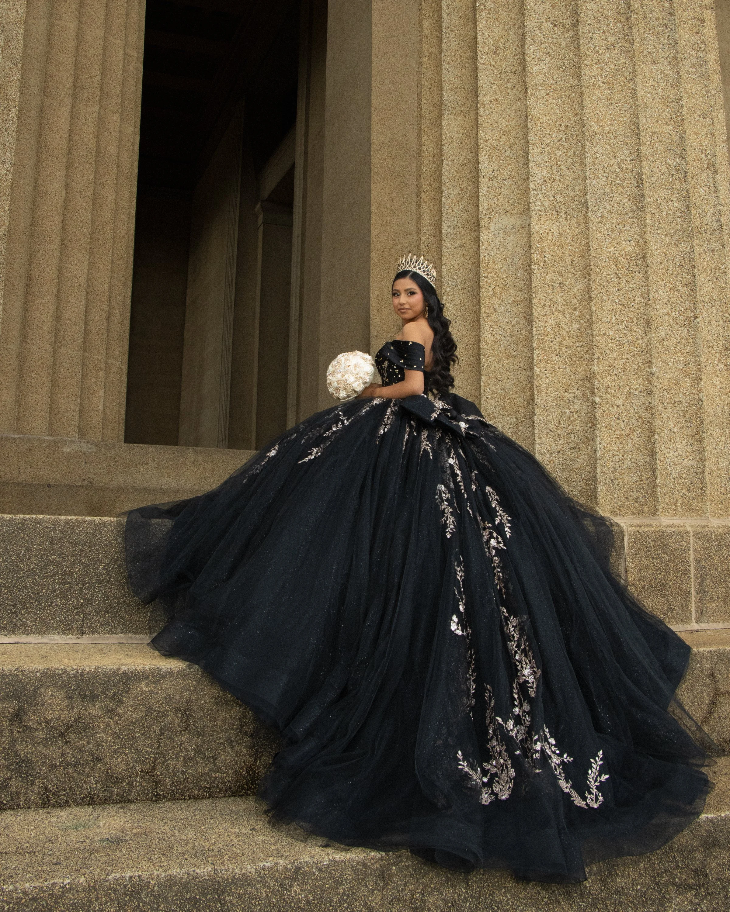A woman in a black, off-the-shoulder ball gown with intricate silver embroidery, wearing a silver tiara, holding a bouquet of white roses, standing on stone steps in front of a textured stone building.