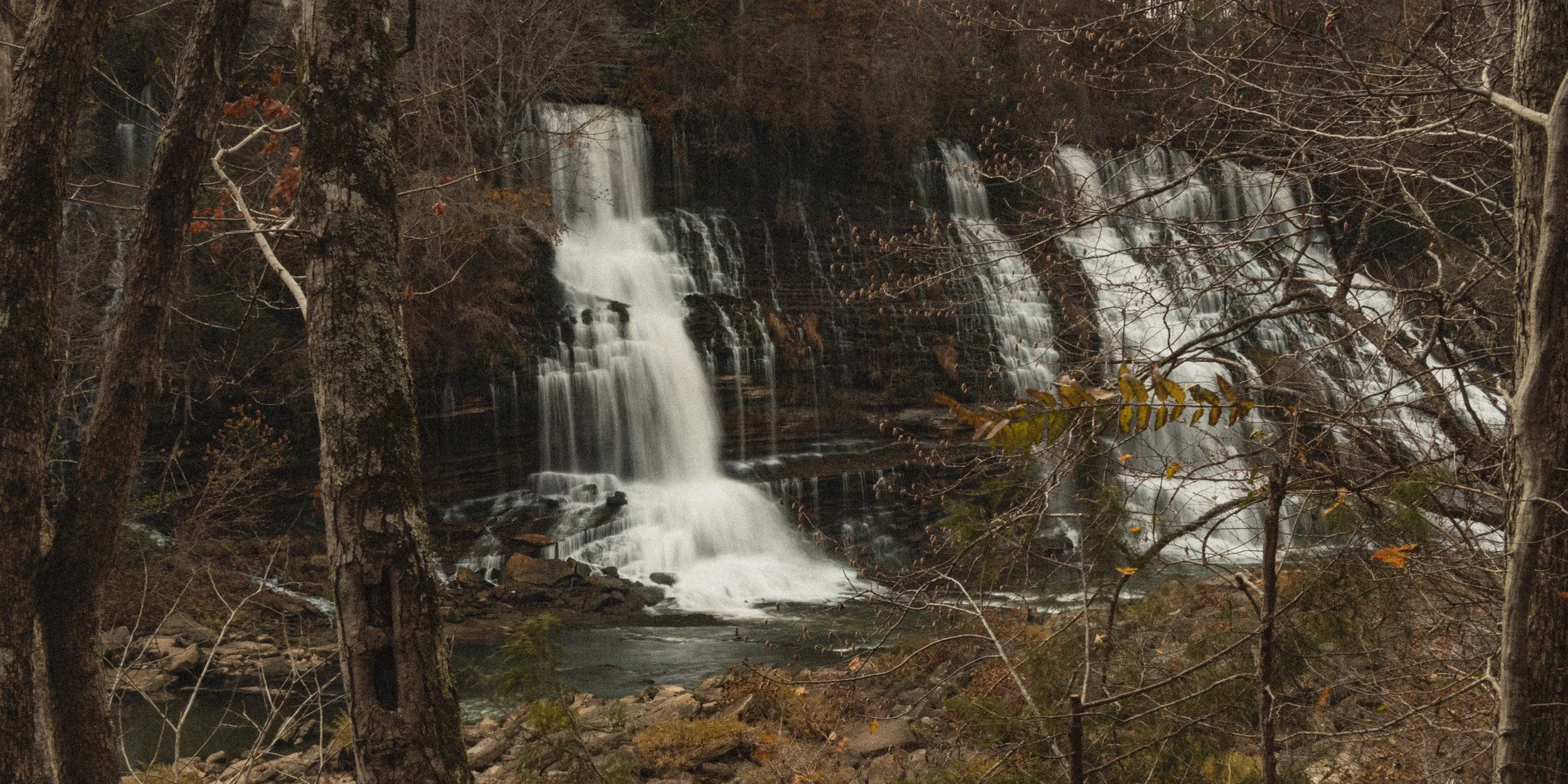 A waterfall cascading down rocks in a wooded area during late autumn or early winter, with leafless trees and sparse foliage.