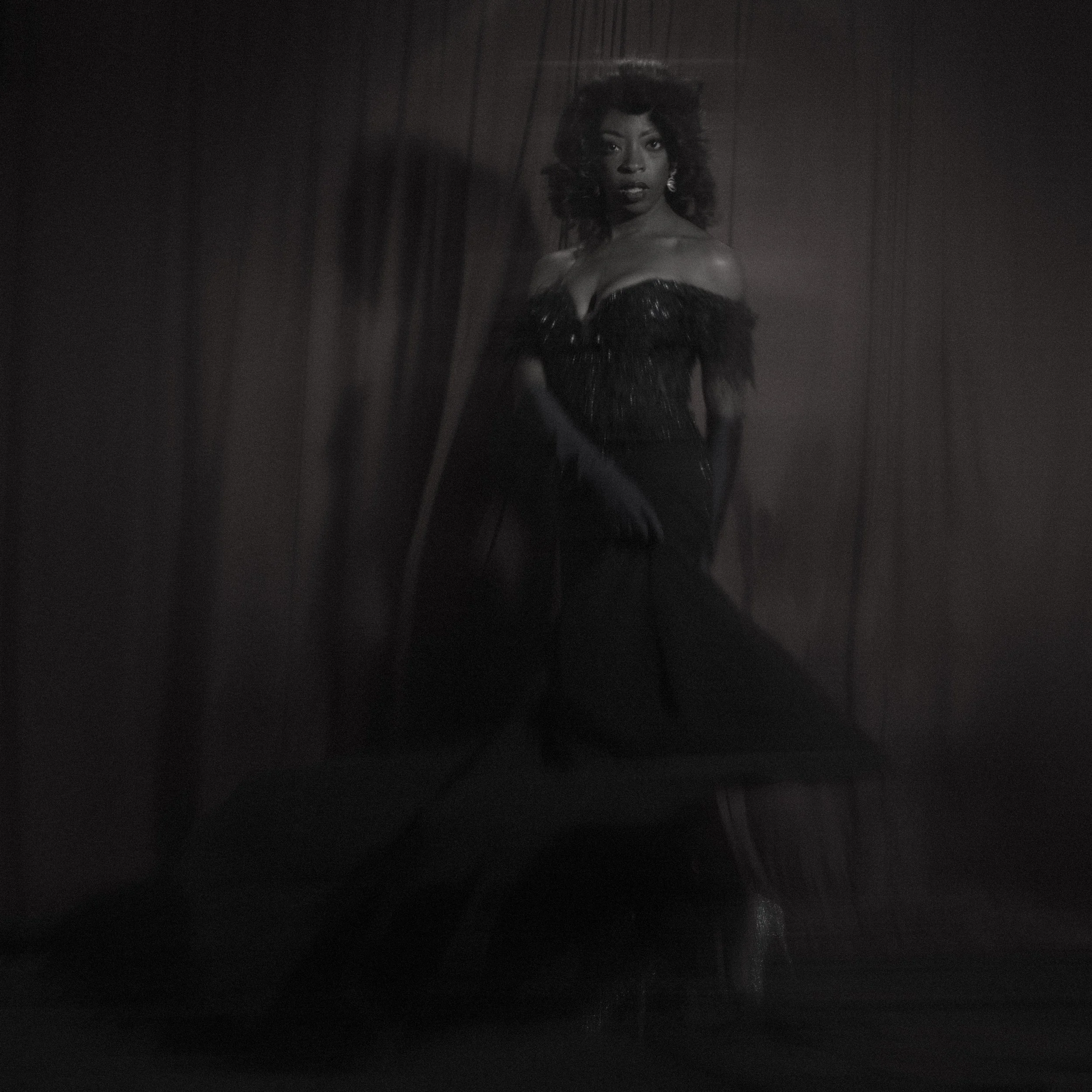 Black and white photo of a woman in a glamorous off-the-shoulder dress, posing against dark curtains.