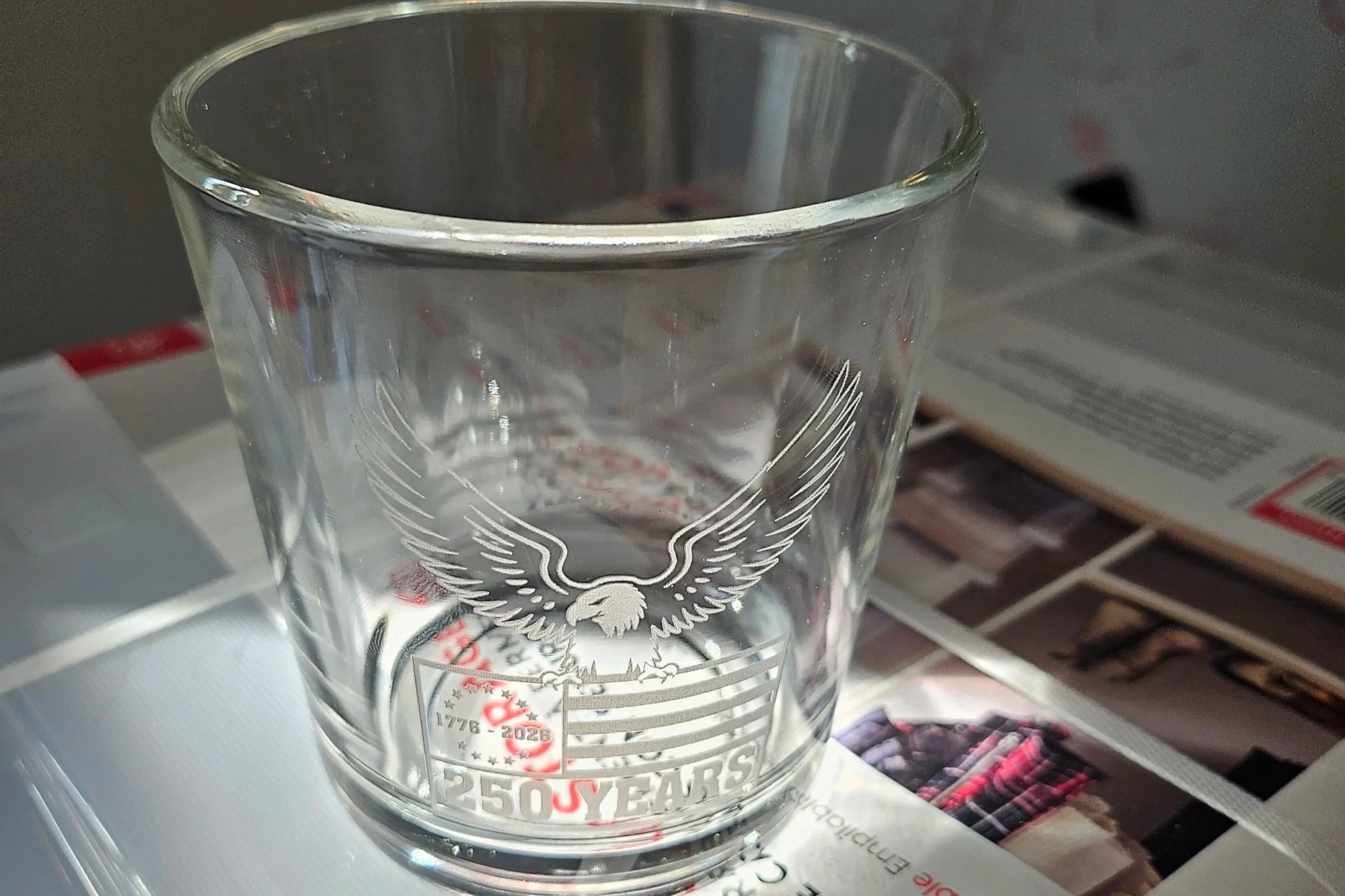 Clear glass tumbler with an etched design of an eagle and an American flag, celebrating 250 years since 1776, with the years 1776-2026 and the phrase "250 YEARS" on a table with magazines or brochures around it.