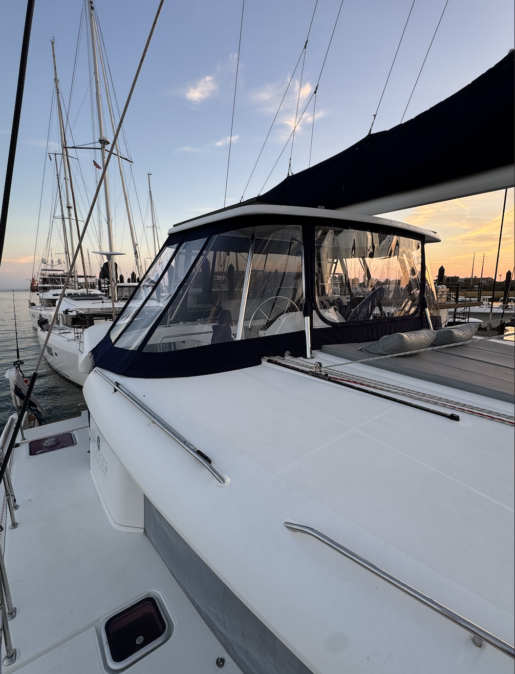 A close-up view of a sailboat docked at a marina during sunset, with other boats visible in the background and the sky partly cloudy.