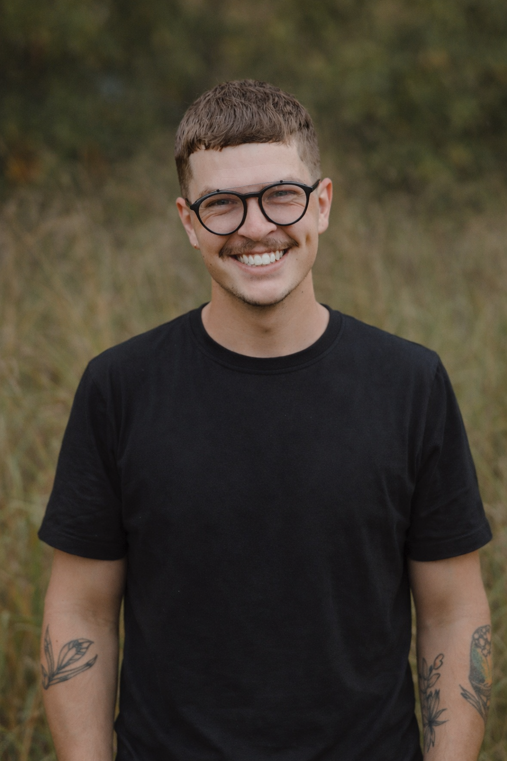 A young man with short brown hair, wearing glasses and a black T-shirt, smiling outdoors in a grassy area.