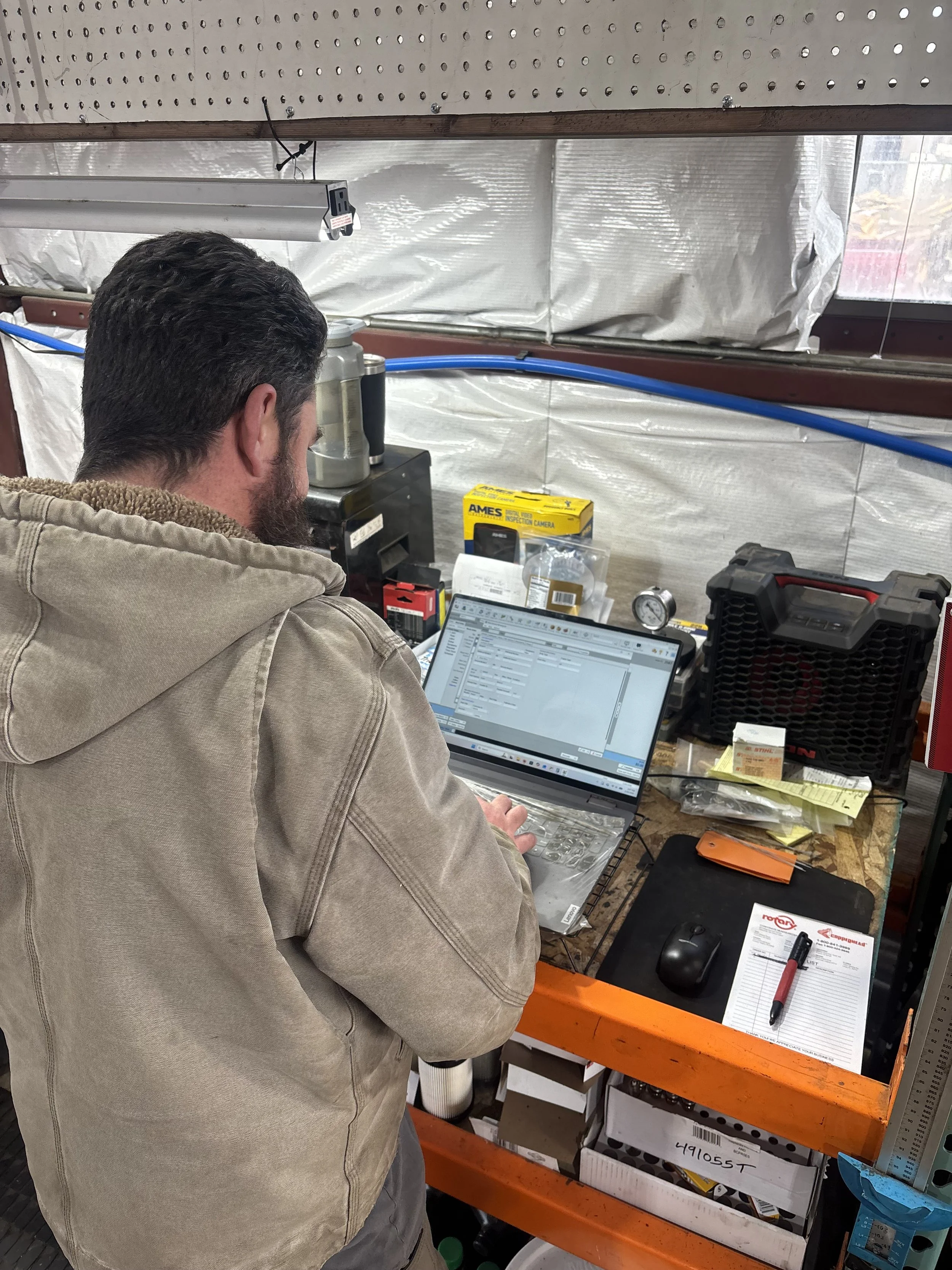 A man with dark hair and a beard wearing a tan hoodie working on a laptop at a cluttered workbench inside a garage or workshop, with various tools and equipment around.