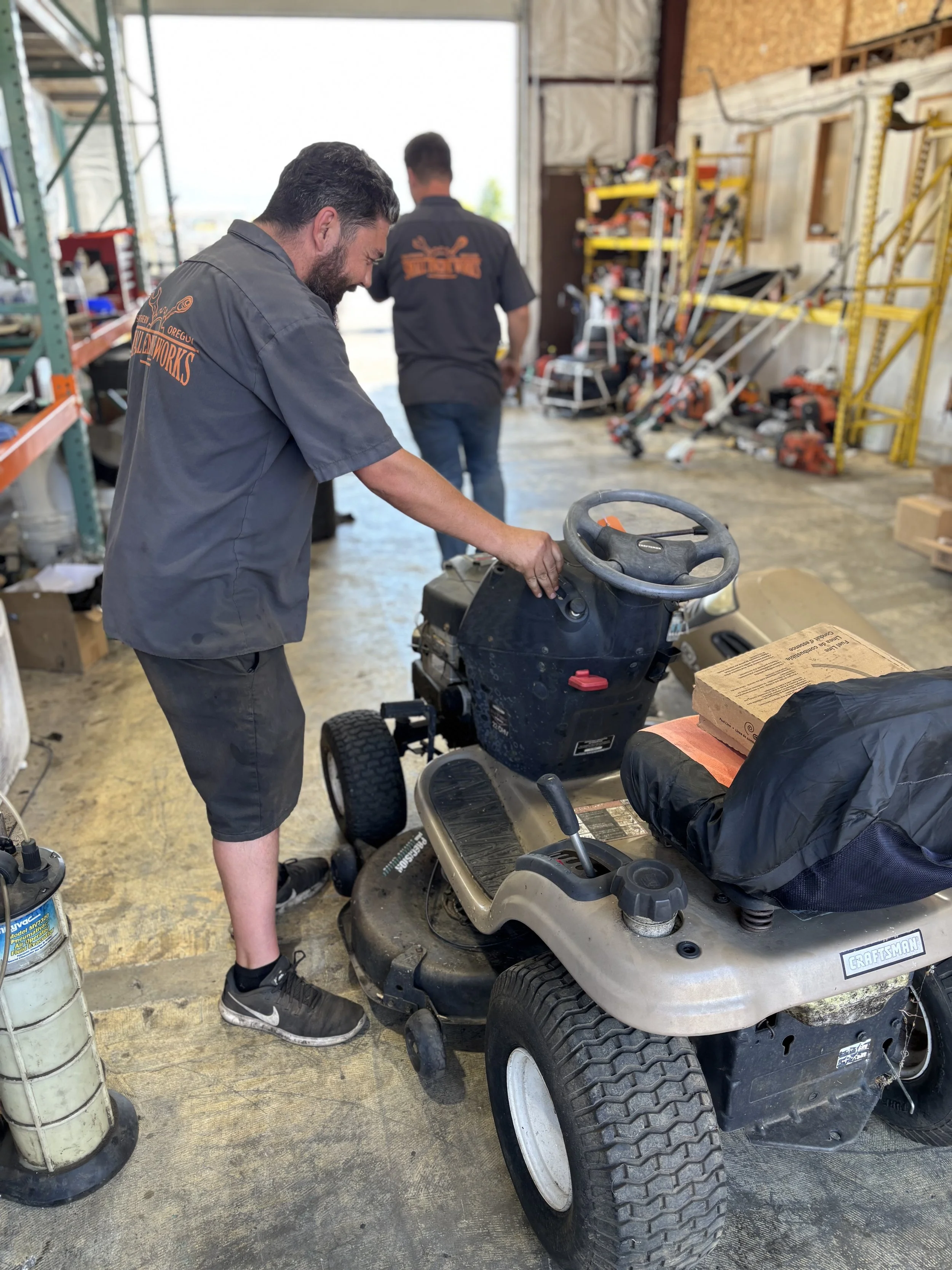 A man standing on a jewelry mower inside a workshop, with two workers in the background and various tools and equipment on shelves.