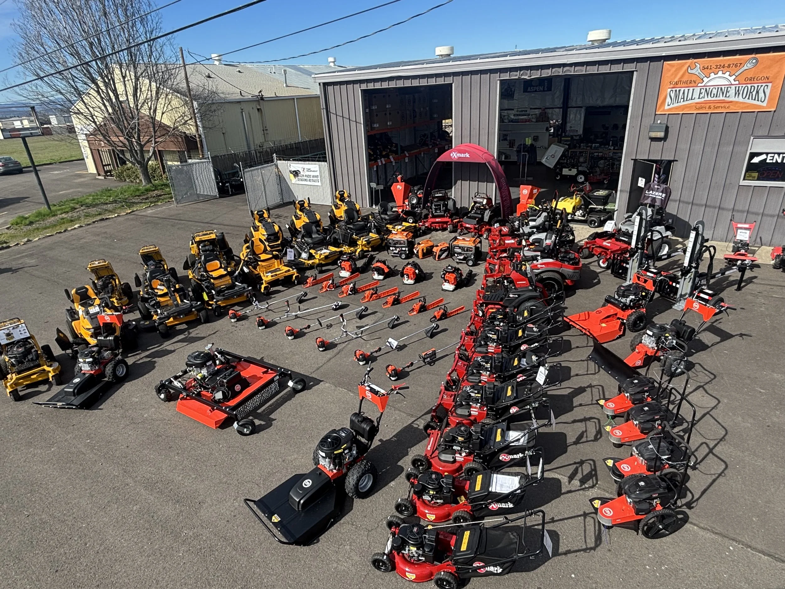 Outdoor display of various lawn equipment including riding mowers, push mowers, and leaf blowers outside a hardware store named Small Engine Works with a grey building and orange signage.