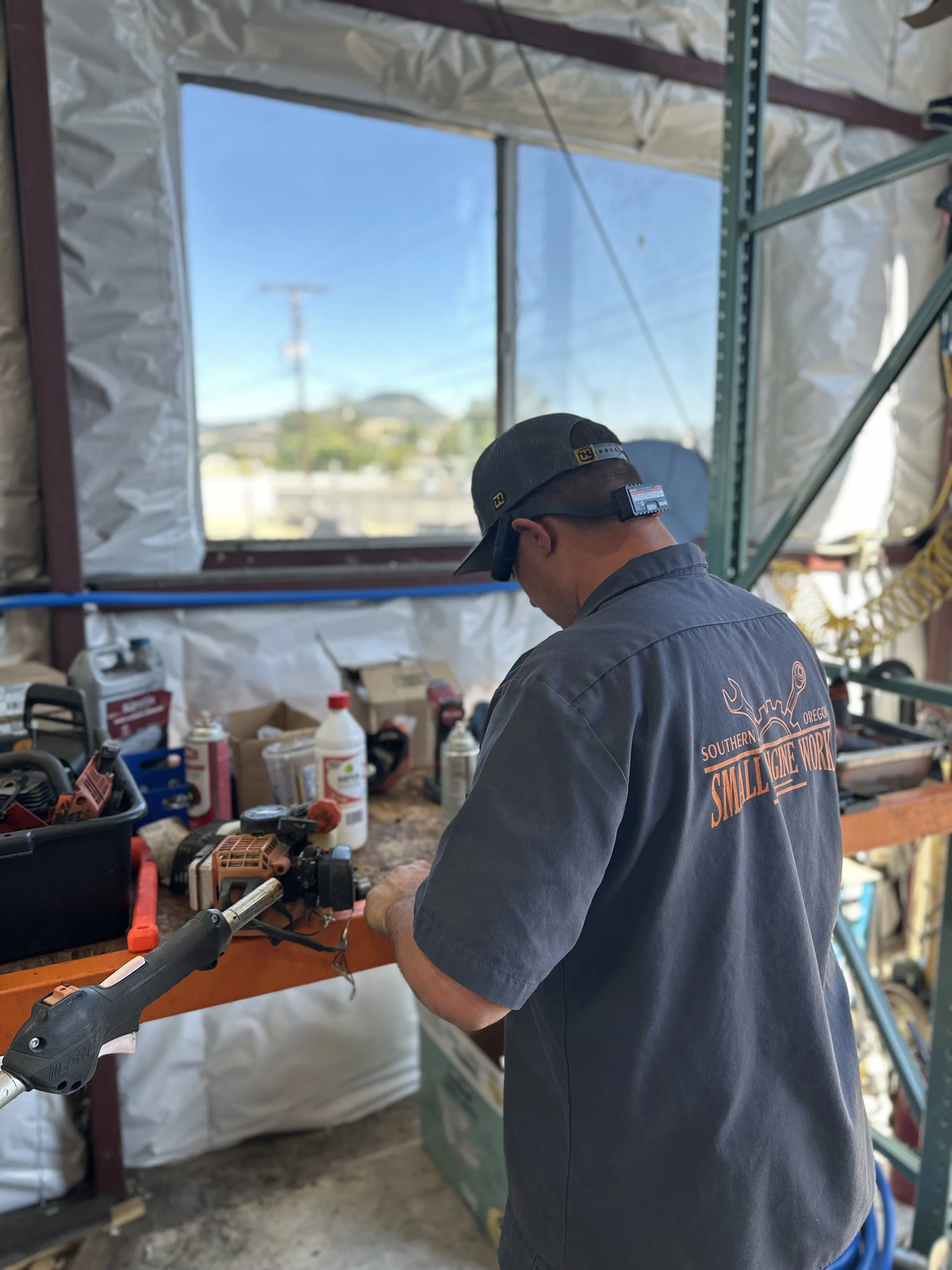 A man wearing a dark gray shirt with orange lettering on the back and a black baseball cap with a headlamp, working at a cluttered workbench inside a garage or workshop, with tools and supplies around him and a large window in the background showing a clear blue sky and some houses.