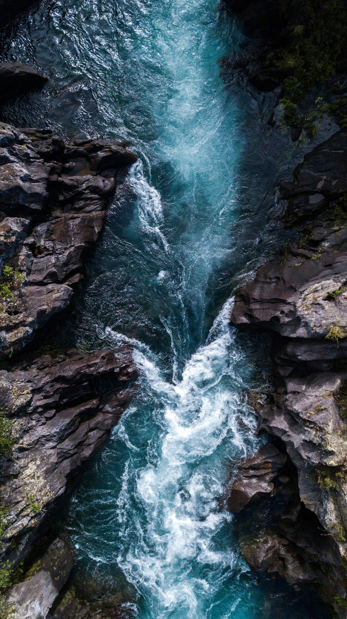 Aerial view of a narrow, rocky mountain stream with rushing, blue water flowing through a canyon surrounded by dark rocks and sparse greenery.