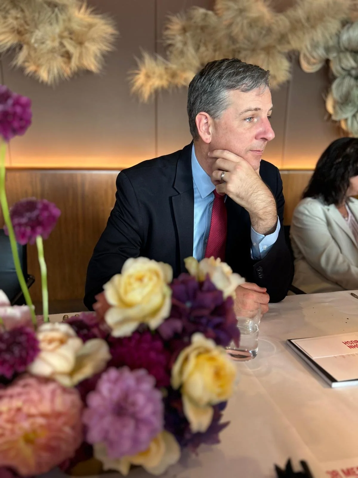 A man in a suit with a red tie sitting at a table decorated with flowers, in a restaurant or conference setting, appearing to listen attentively.