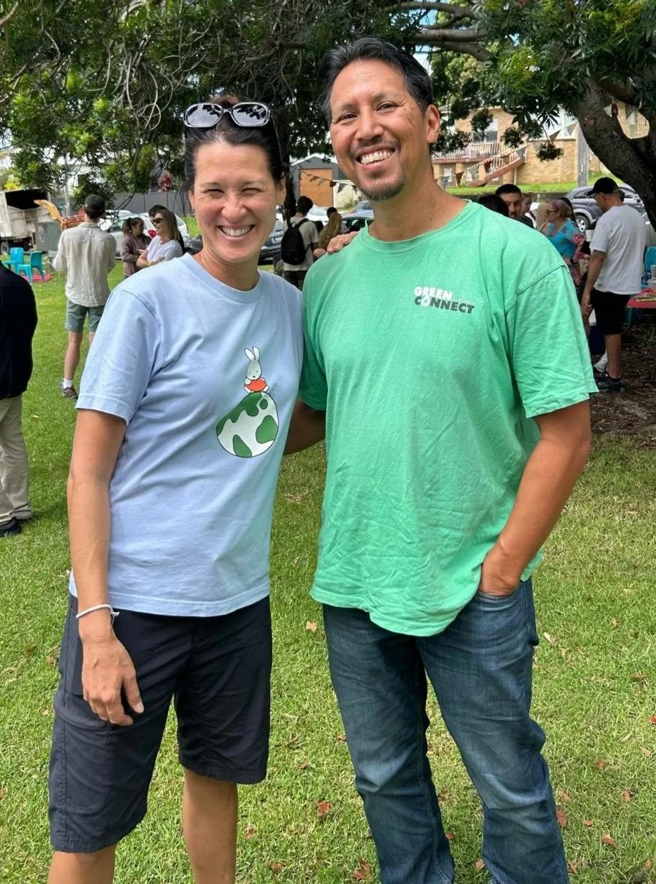 Two people, a woman and a man, smiling at an outdoor event, with other people in the background, standing under a tree, on a grassy area.