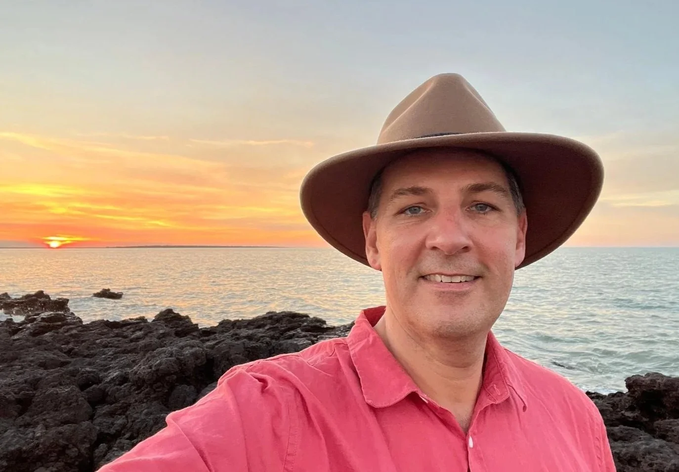 Man taking selfie at sunset on rocky shoreline, wearing a wide-brimmed hat and red shirt.