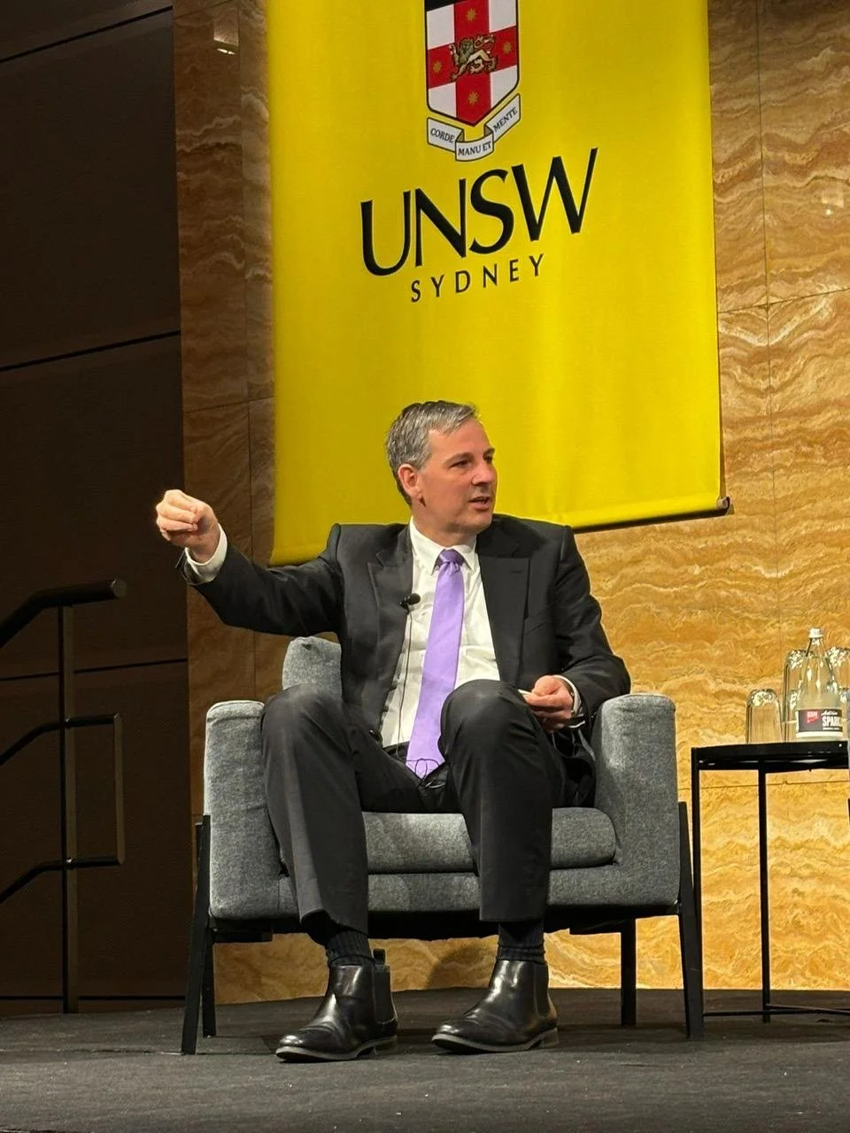 A man in a suit and purple tie sitting in a gray armchair on stage at UNSW Sydney, gesturing with his right arm, with a yellow UNSW banner hanging behind him.