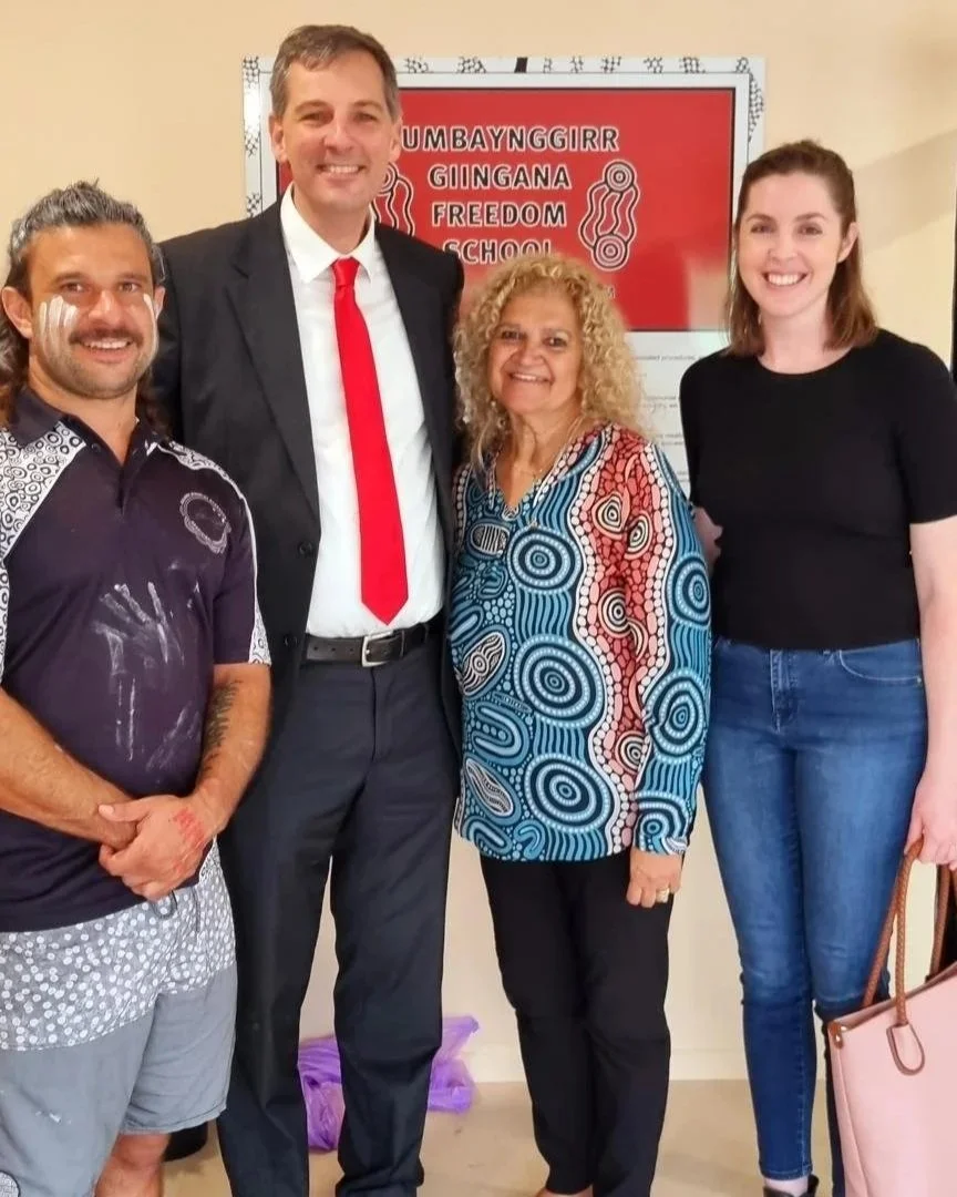 Four people standing and smiling in front of a sign that reads 'GUMBAYNGGIRR GINGANA FREEDOM SCHOOL' One Aboriginal man in in ochre and casual clothes, one European man in a suit and tie, and an Aboriginal woman and a European woman in casual clothes