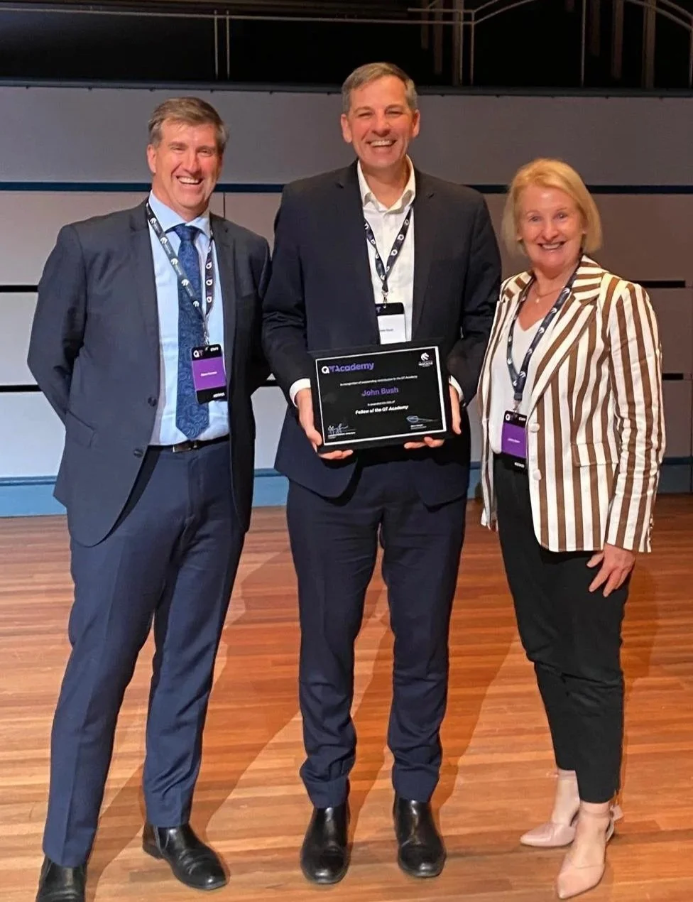 Three professionally dressed people standing on a wooden floor at an event, smiling, with one holding a plaque.