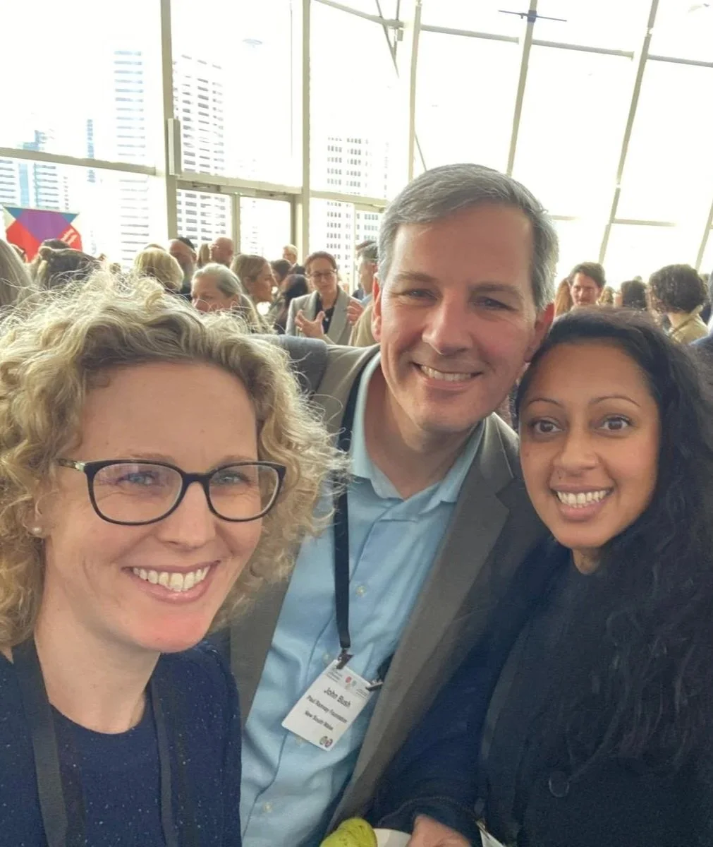 Three people smiling for a selfie at a crowded indoor event with large windows and city skyline in the background.