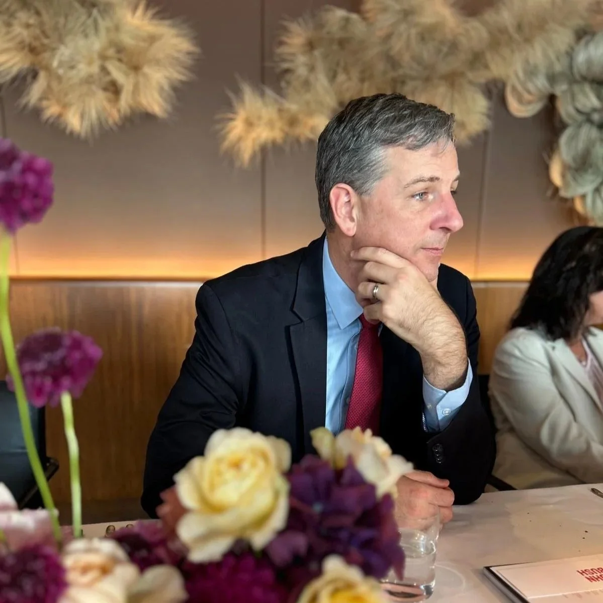 A man in a navy suit, blue shirt, and red tie sitting at a table during a formal event, resting his chin on his hand, with a thoughtful expression. Flowers in purple, cream, and yellow are visible in the foreground, and a woman in a light-colored blazer is partially visible next to him. Decorative wall art and soft lighting are in the background.