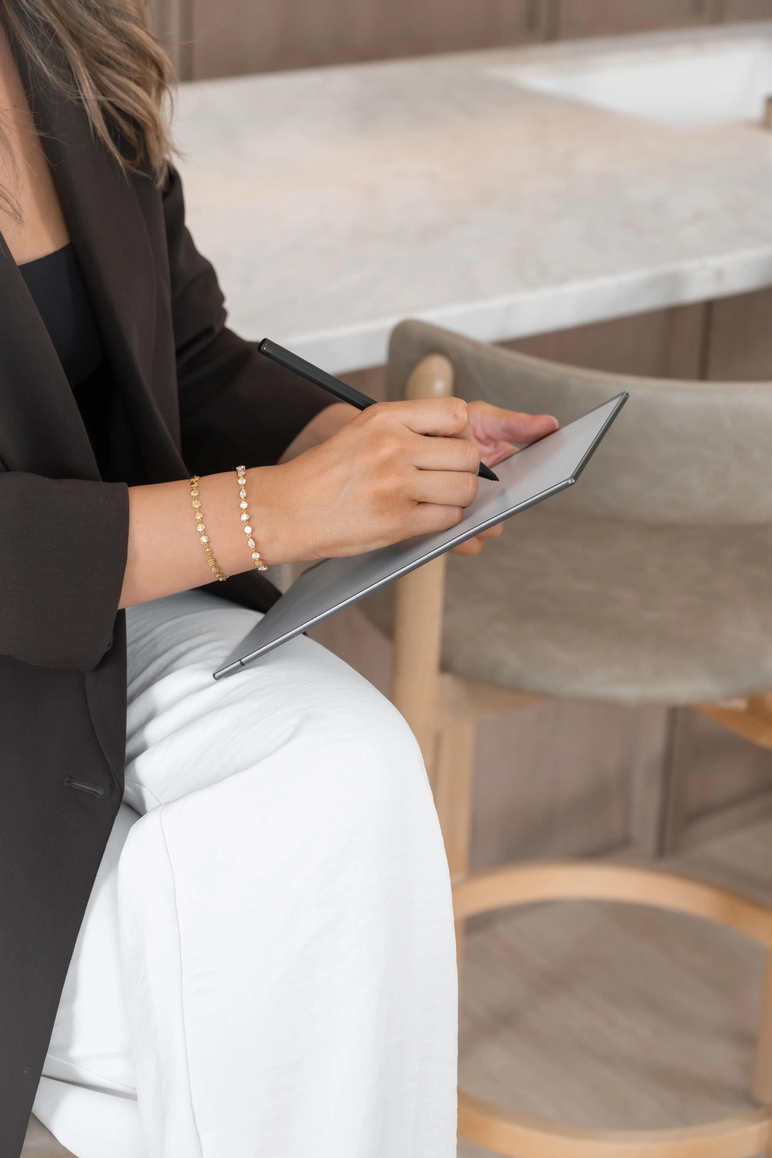 A woman sitting on a chair, wearing a black blazer and white pants, writing on a gray tablet with a stylus in a modern indoor space.