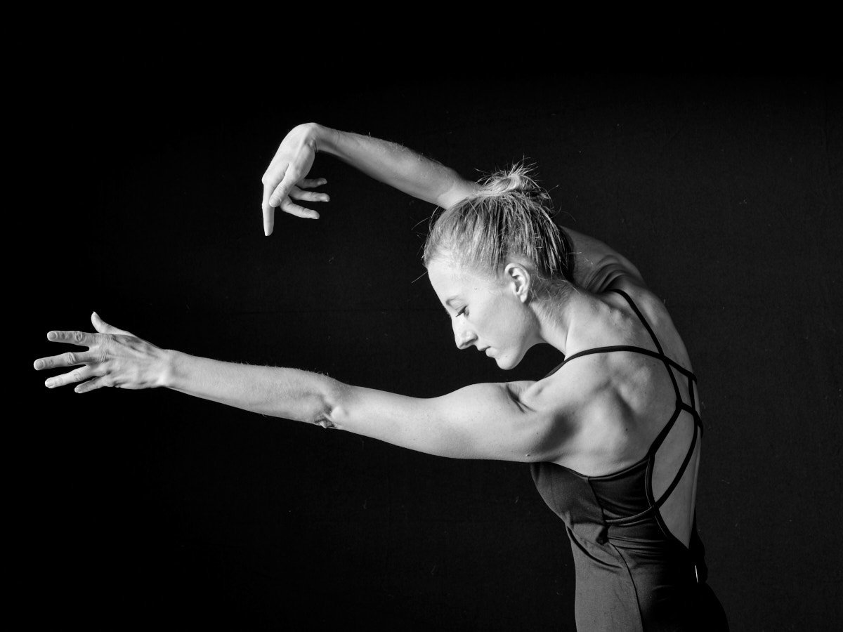 Michaela Ellingson in a strappy black top stretching with one arm raised above her head and the other extended forward against a black background.