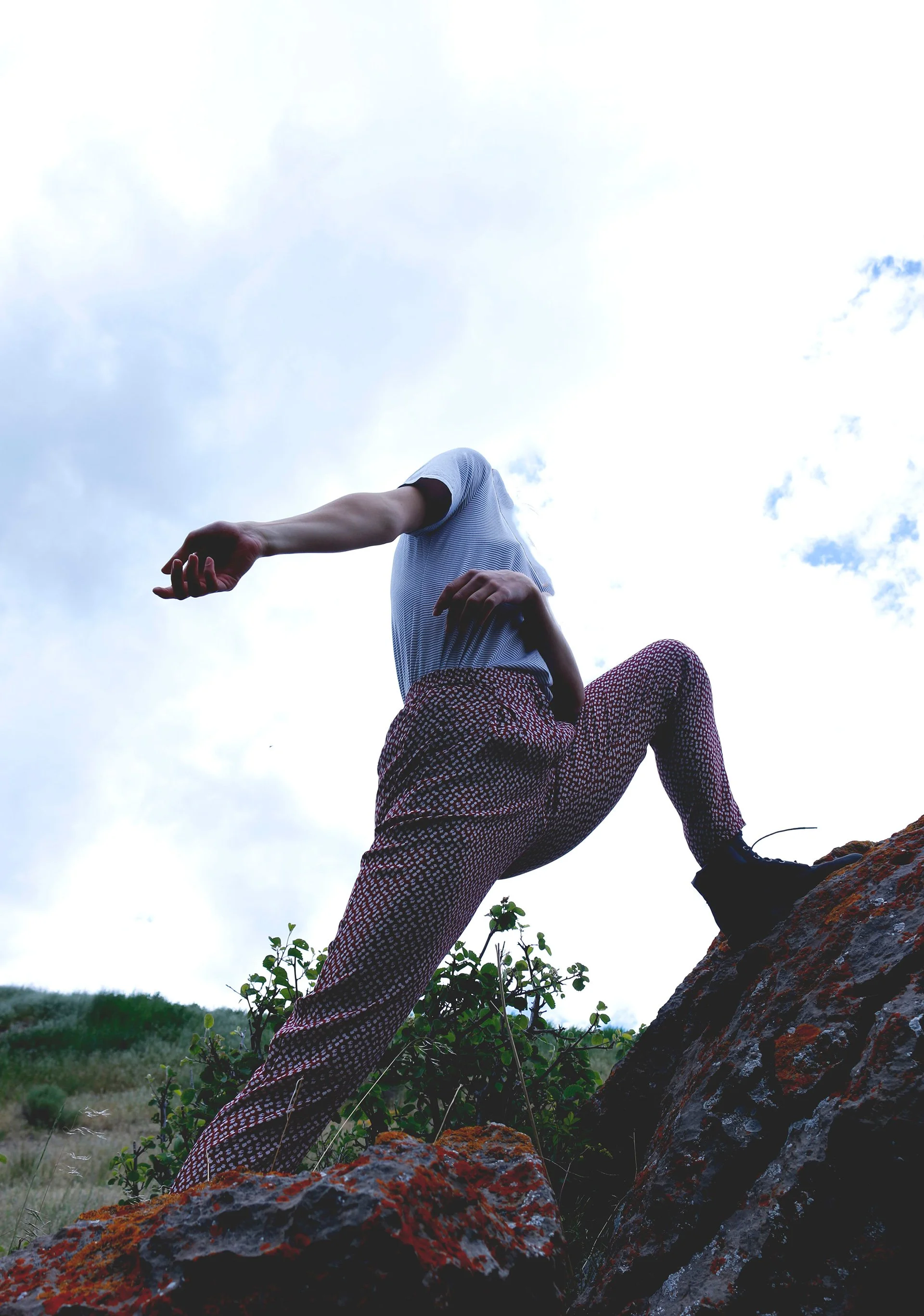 A person is climbing on a rocky terrain outdoors, with the sky and clouds in the background. The person is wearing patterned pants and a striped shirt, with their face not visible.