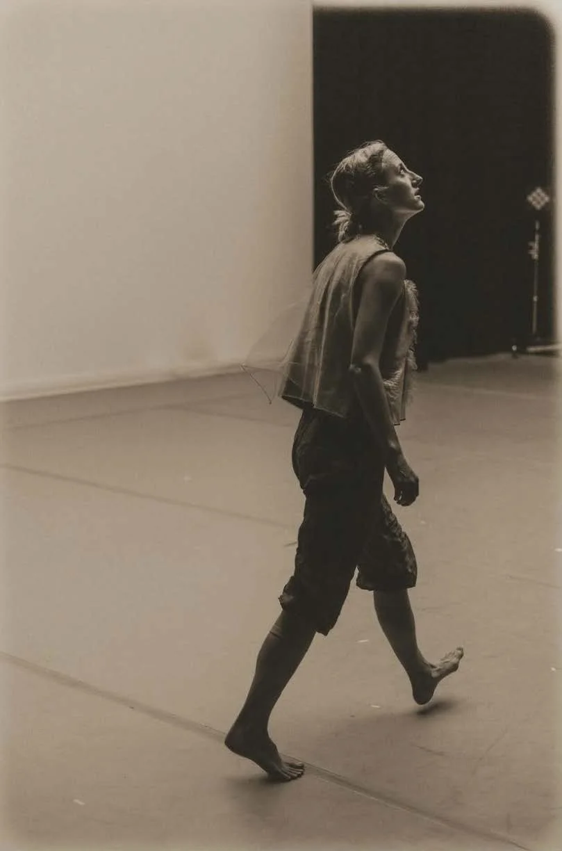 Michaela Ellingson in casual dance clothing walking barefoot across a spacious, empty room with plain walls, looking upward, illuminated by soft lighting.