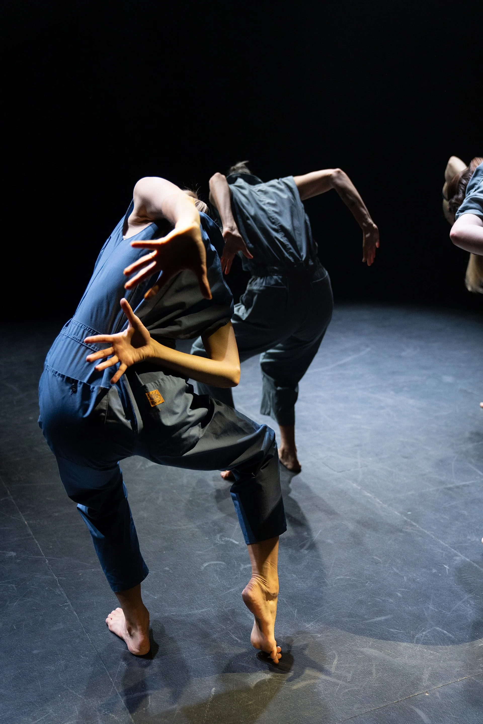 Three performers in dark clothing are dancing on stage against a black background. Their poses are dynamic and expressive, with bent knees, arched backs, and extended arms, showcasing a modern or contemporary dance style.