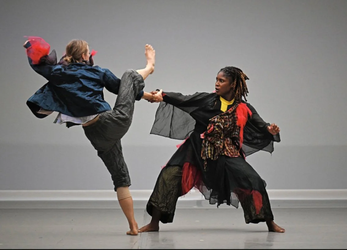 Michaela Ellingson engaged in a dance performance on stage, with one woman lifting her leg high while the other holds her arm in a dance pose.