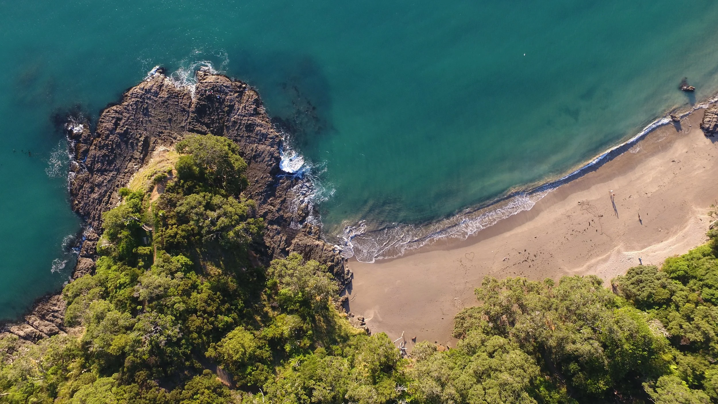 An aerial view of a beach with a rocky outcrop covered in greenery, gentle waves hitting the shore, and a few people walking on the sandy beach.