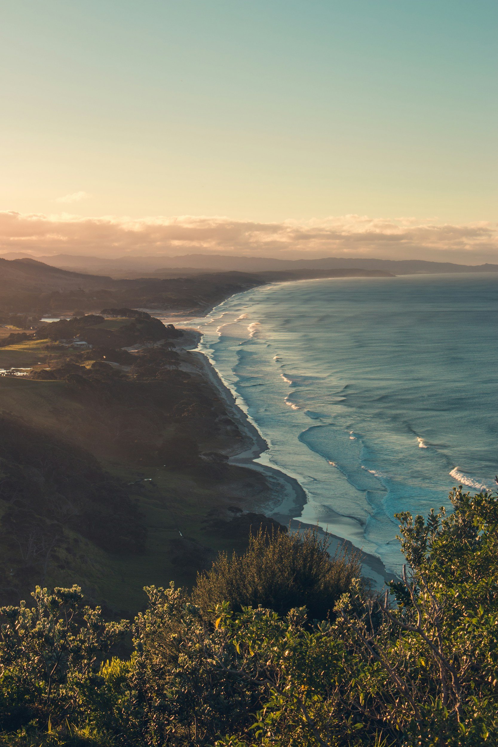 Coastal landscape with rolling hills, a sandy beach, and ocean waves under sunset sky with clouds.
