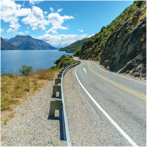 A winding coastal road runs alongside a lake with mountains in the background on a sunny day with a partly cloudy sky.