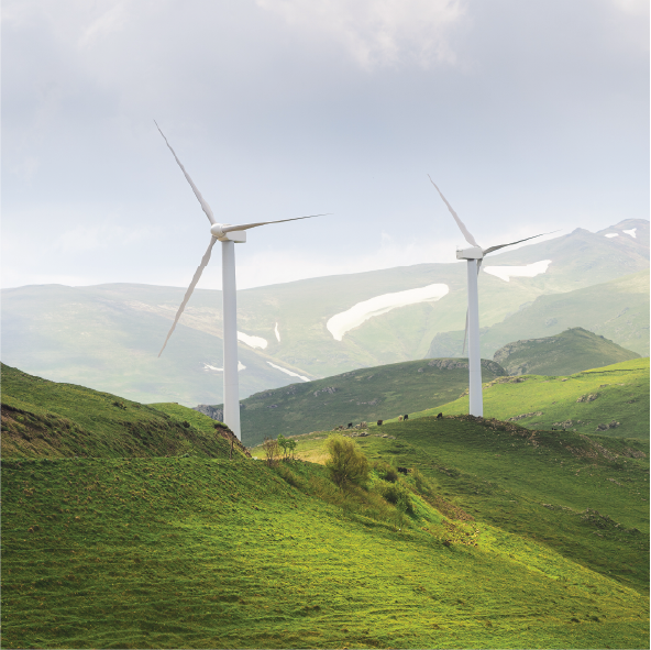 Two wind turbines on green rolling hills in a mountainous landscape with patches of snow.