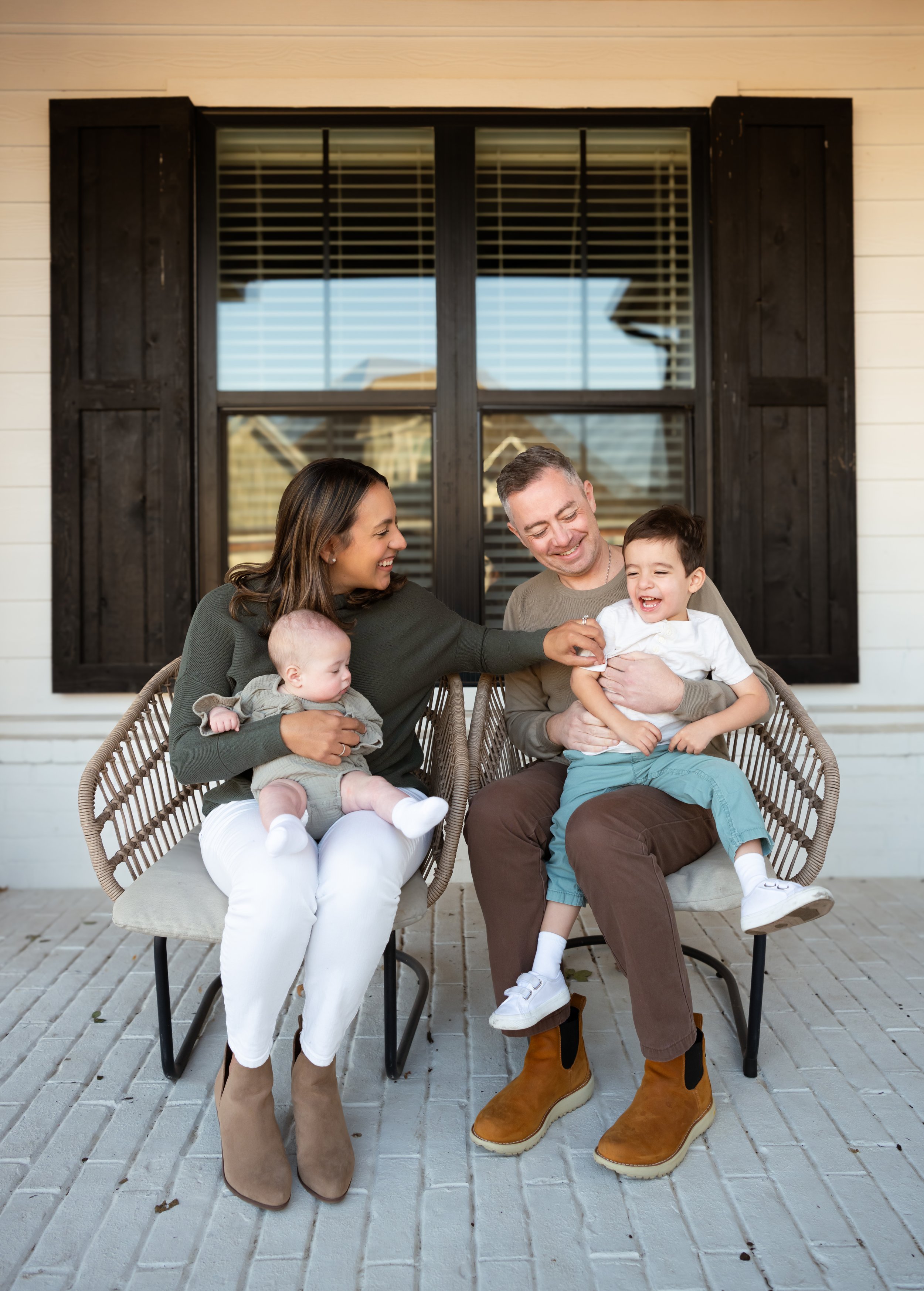 A happy family of four sitting on a porch in front of a house window, smiling and playing together. The parents each hold a young child, and the children are laughing.