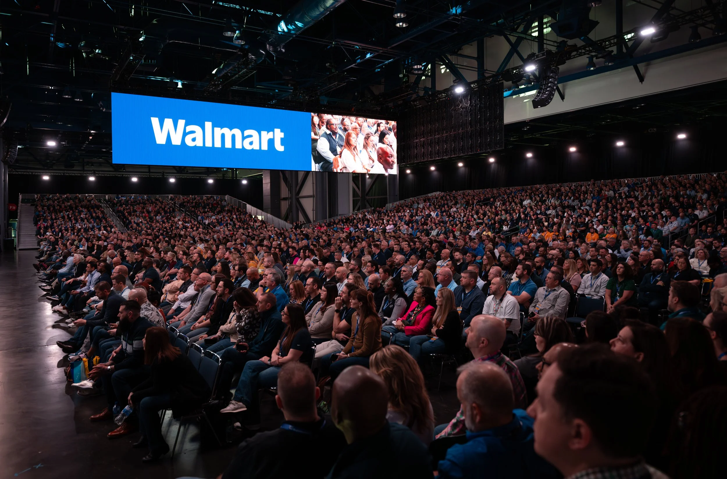 A large audience attending a conference or presentation in a dark auditorium with a big screen displaying the 'Walmart' logo.