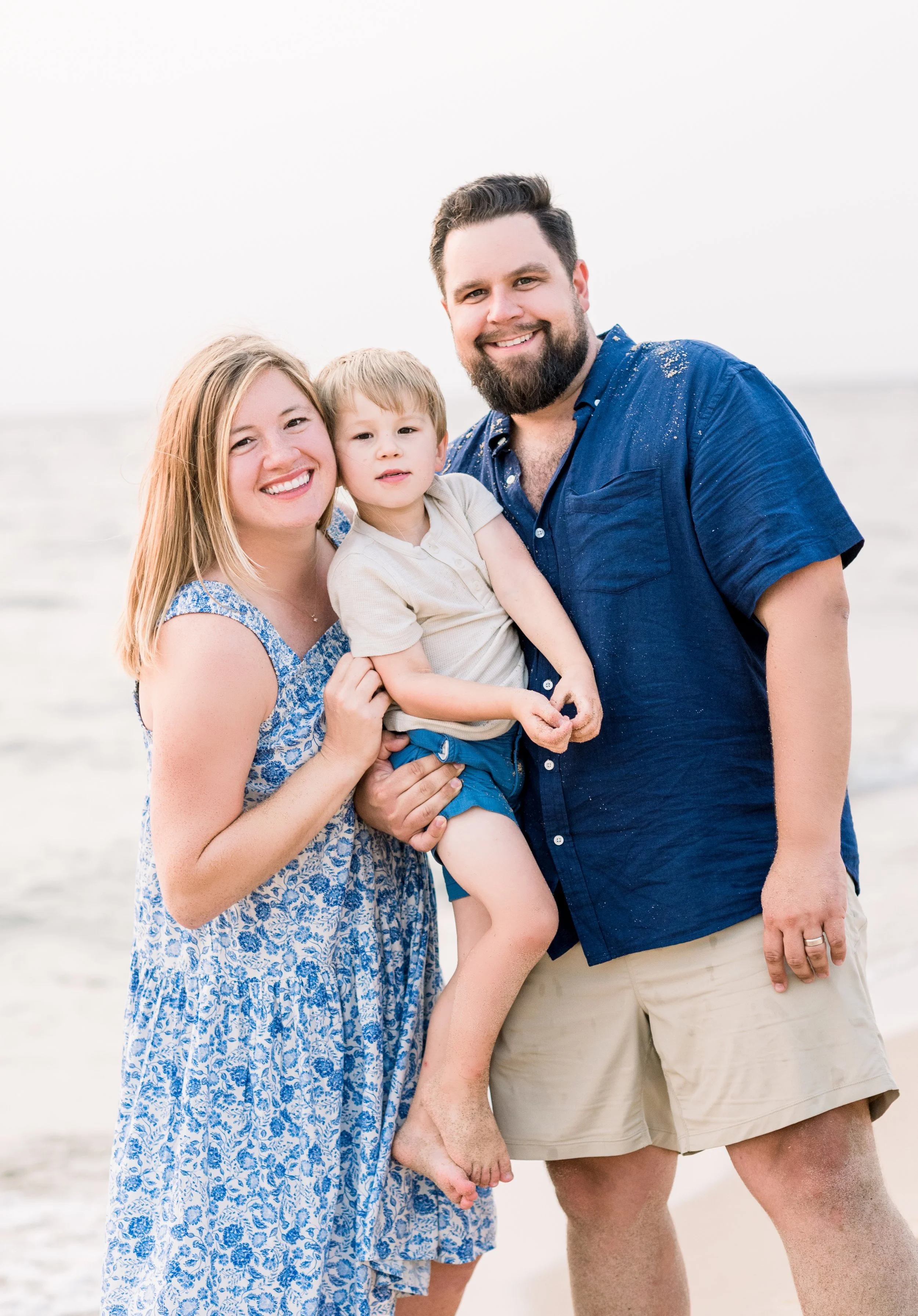 A happy family of three standing on the beach, smiling at the camera with ocean in the background. The woman is wearing a blue patterned dress, the man a blue shirt and tan shorts, and the young boy in a beige shirt and blue shorts.