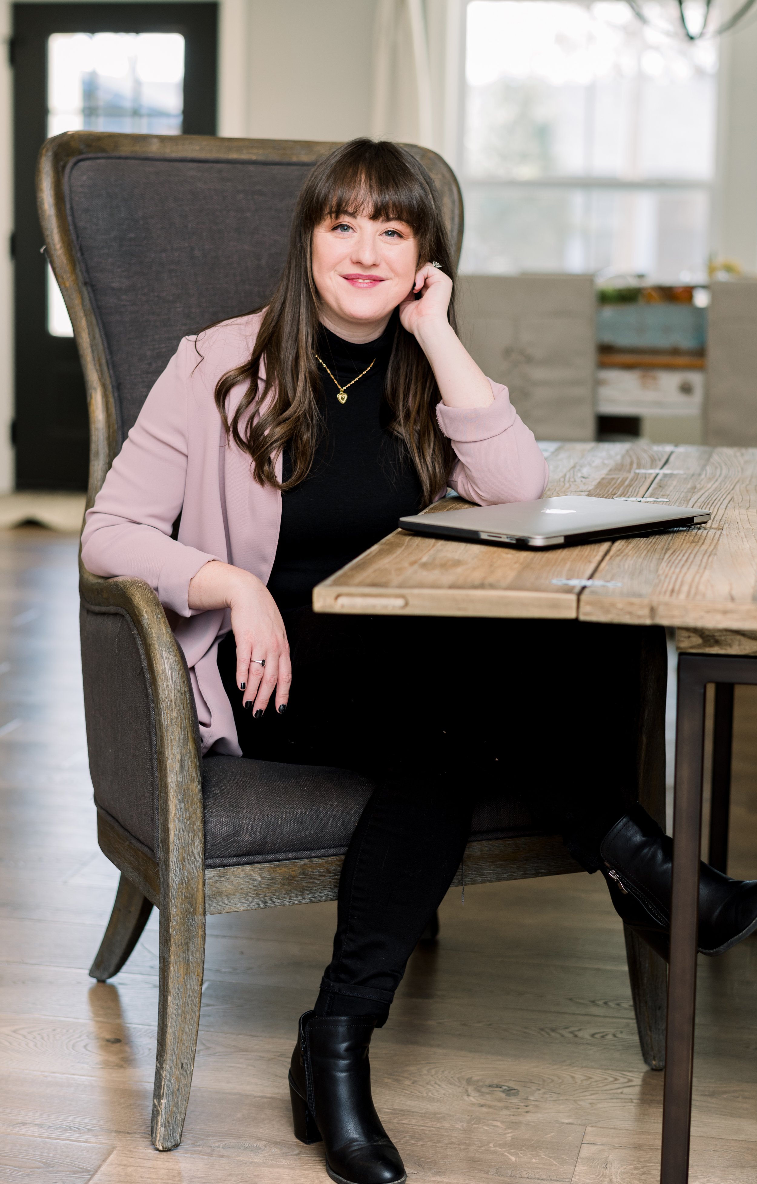 A woman with long brown hair sitting at a wooden table. She is smiling and wearing a black top, a pink blazer, black pants, and black high-heeled boots. There is a closed laptop on the table and a window in the background.