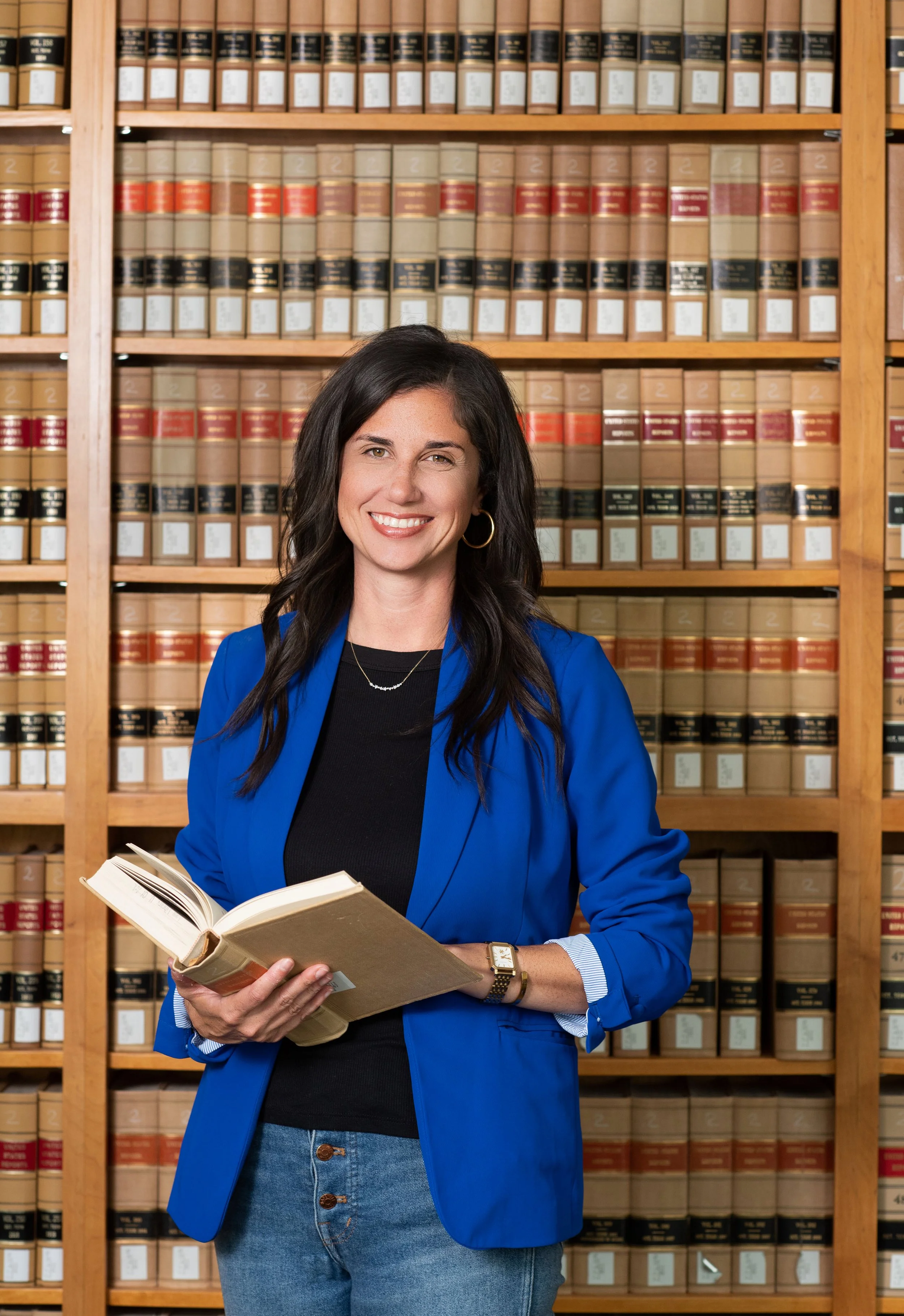 Smiling woman with dark hair, gold hoop earrings, blue blazer, black top, blue jeans, holding open book, standing in front of bookshelf filled with legal books.