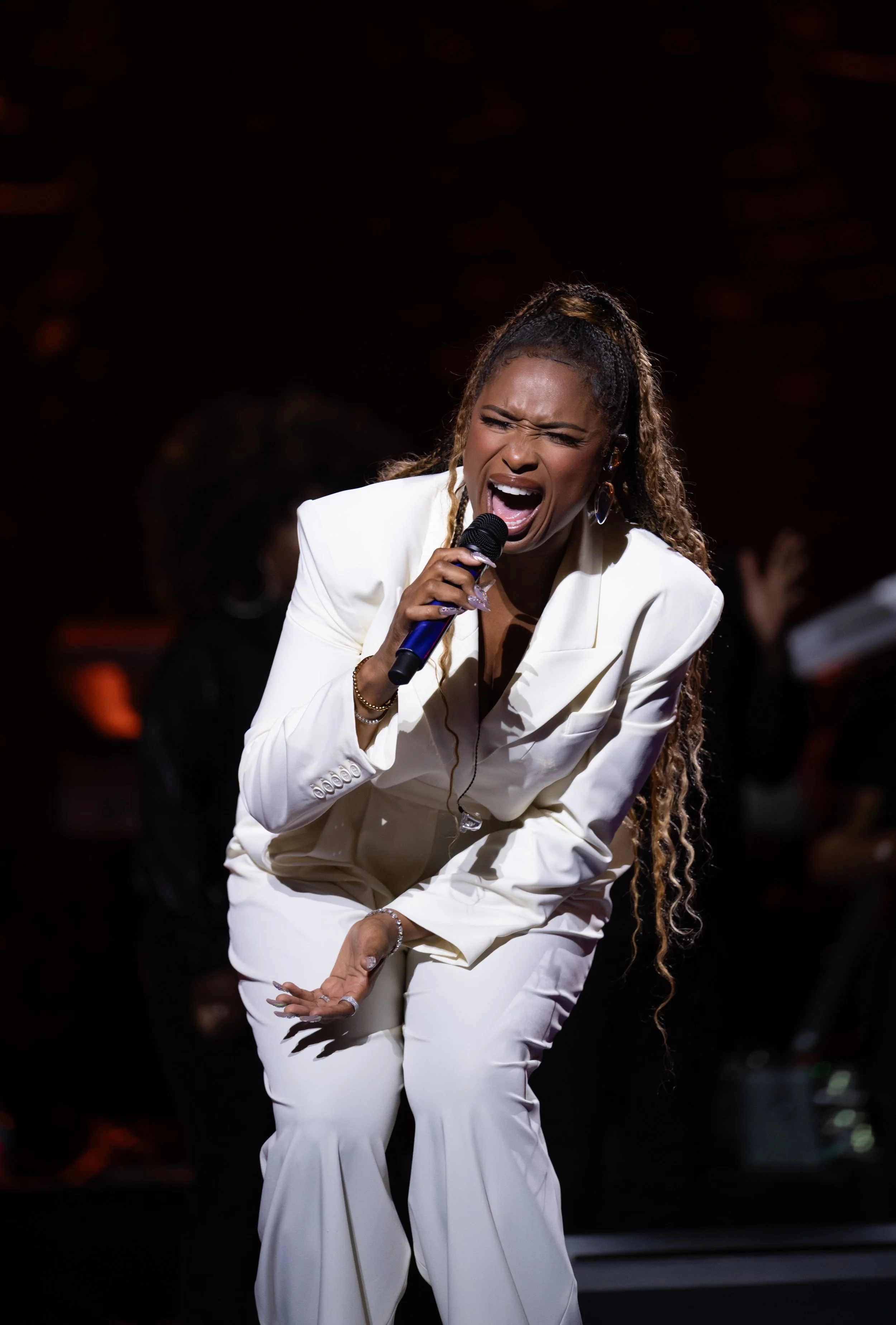Jennifer Hudson dressed in a white suit passionately singing into a microphone on stage, with a dark background and some out-of-focus people.