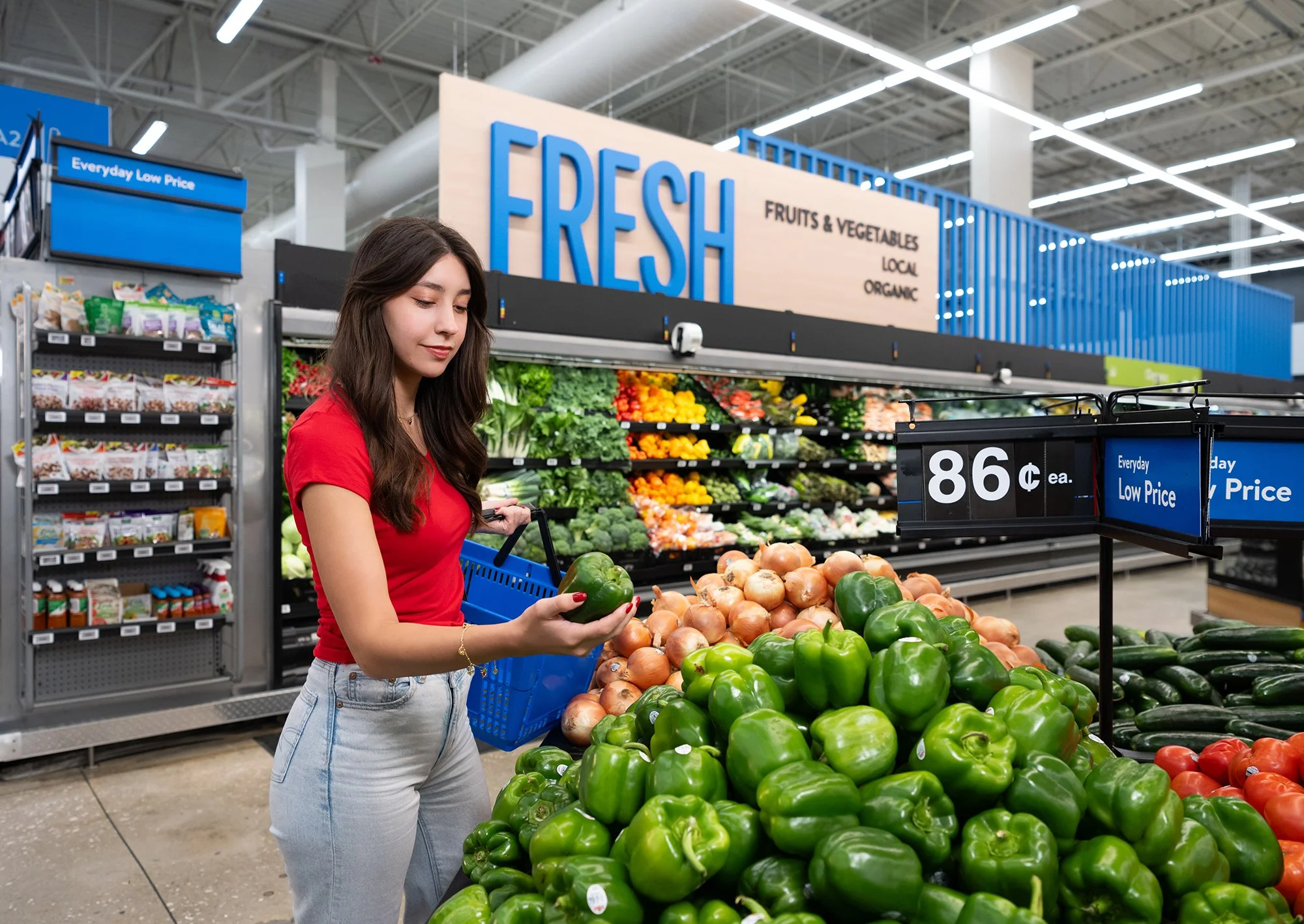 A young woman shopping for produce at a grocery store, selecting a green bell pepper from a display of fresh vegetables.