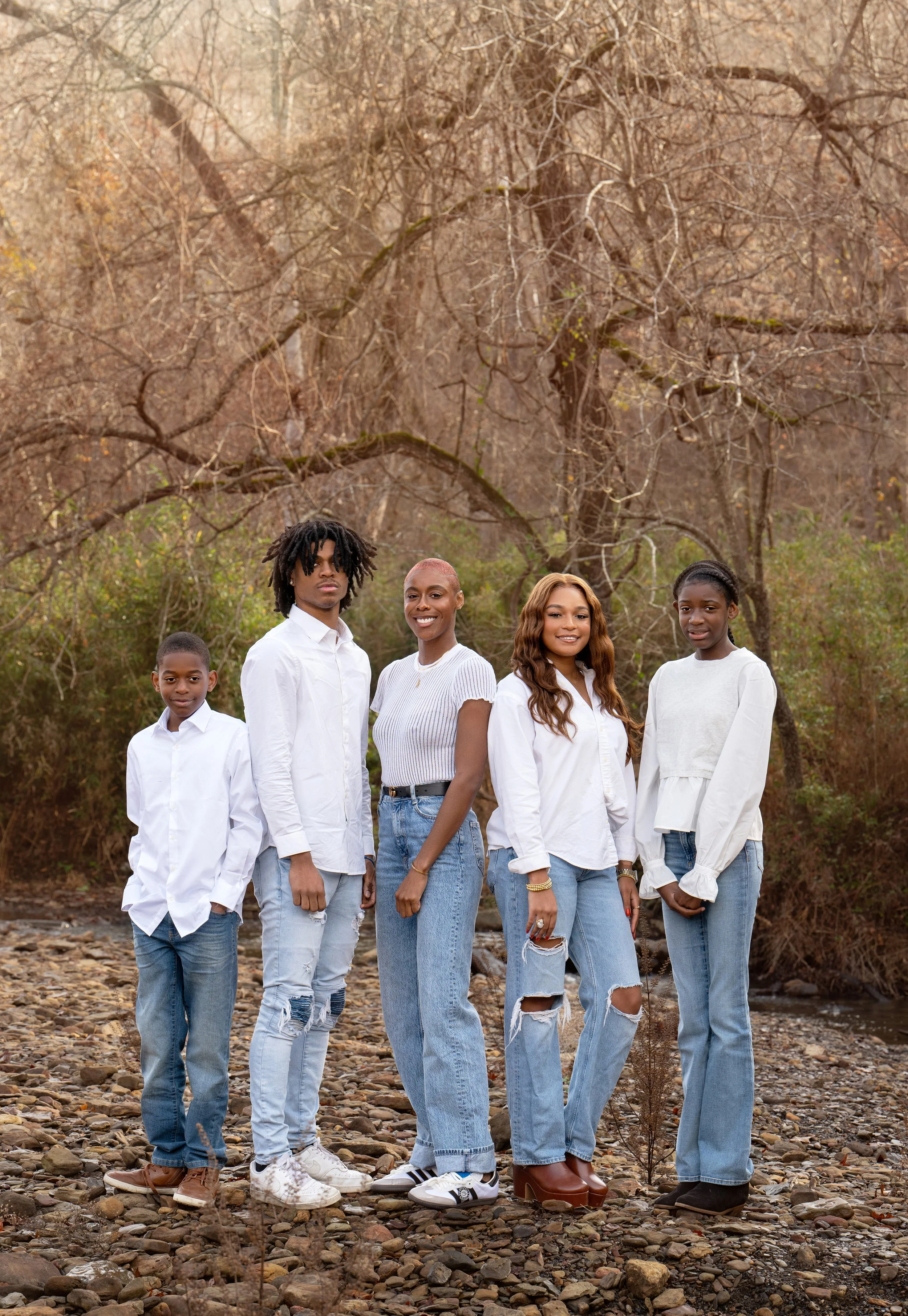 Group of five people standing outdoors on a rocky ground with bare trees in the background, wearing white tops and blue jeans.