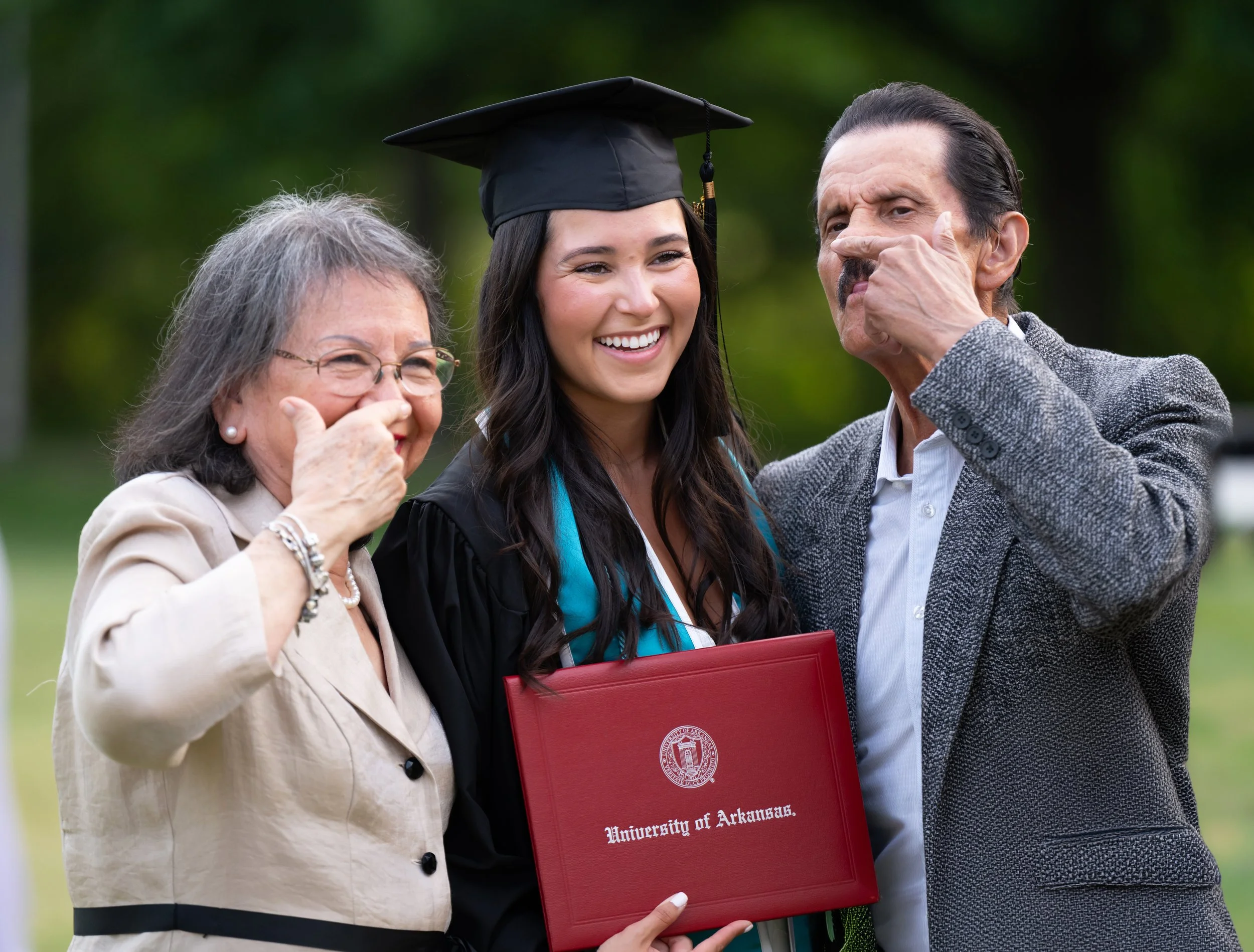 A young woman in a graduation cap and gown holding a diploma, smiling with her parents during an outdoor graduation celebration.