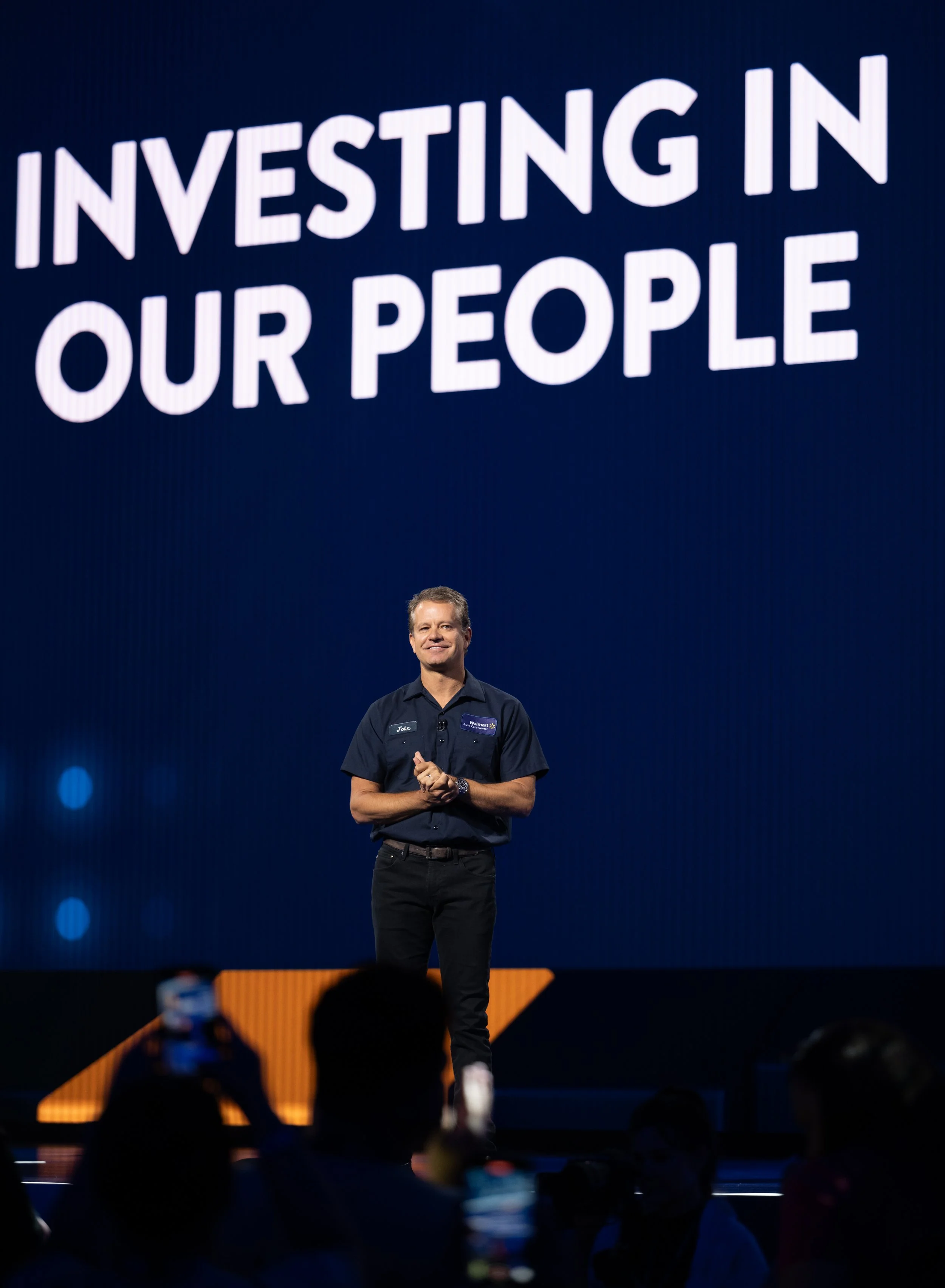 A man standing on stage in front of a large screen that reads 'Investing in Our People', with an audience taking photos of him.