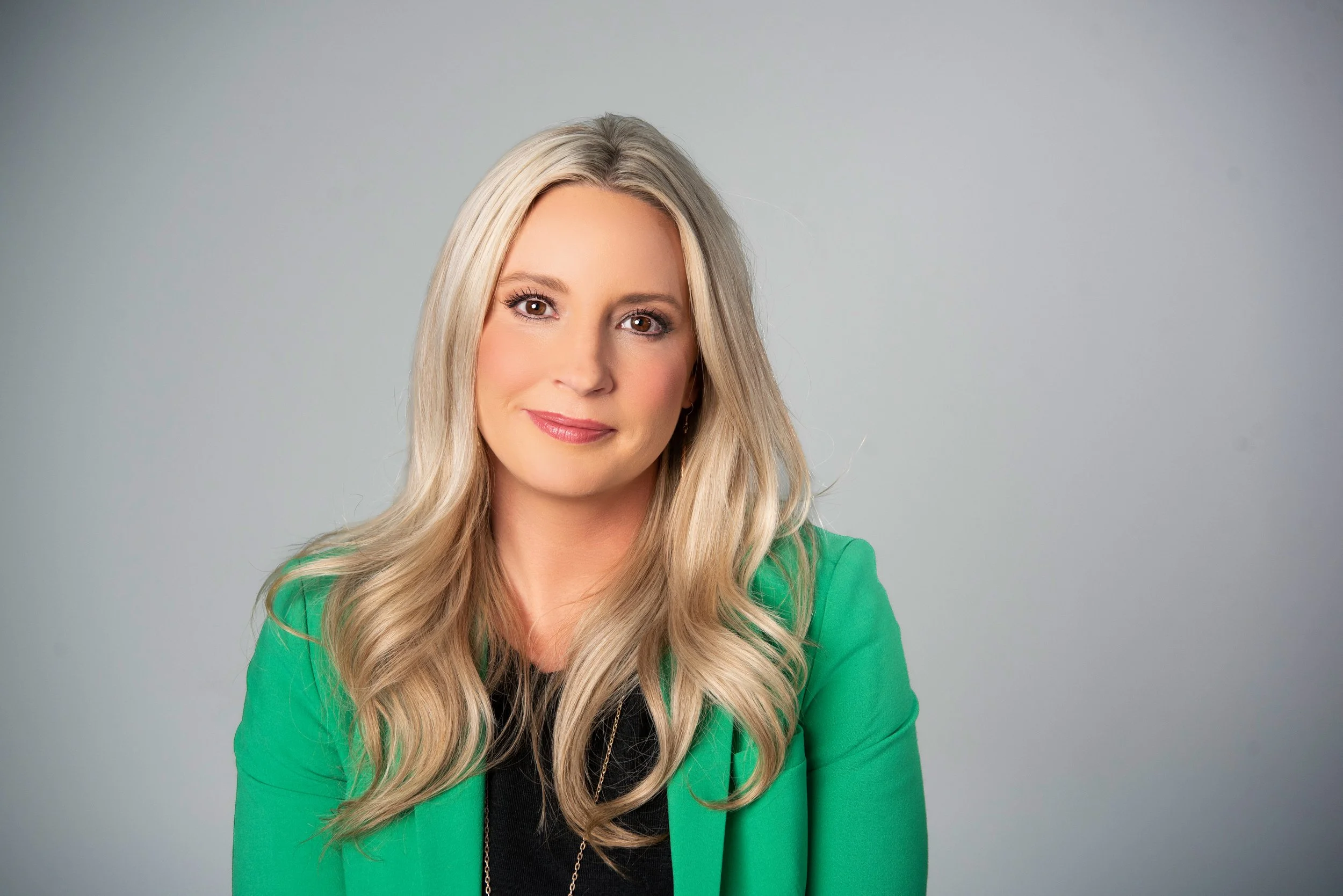 A woman with long blonde hair, wearing a green blazer over a black top, sitting against a plain gray background.