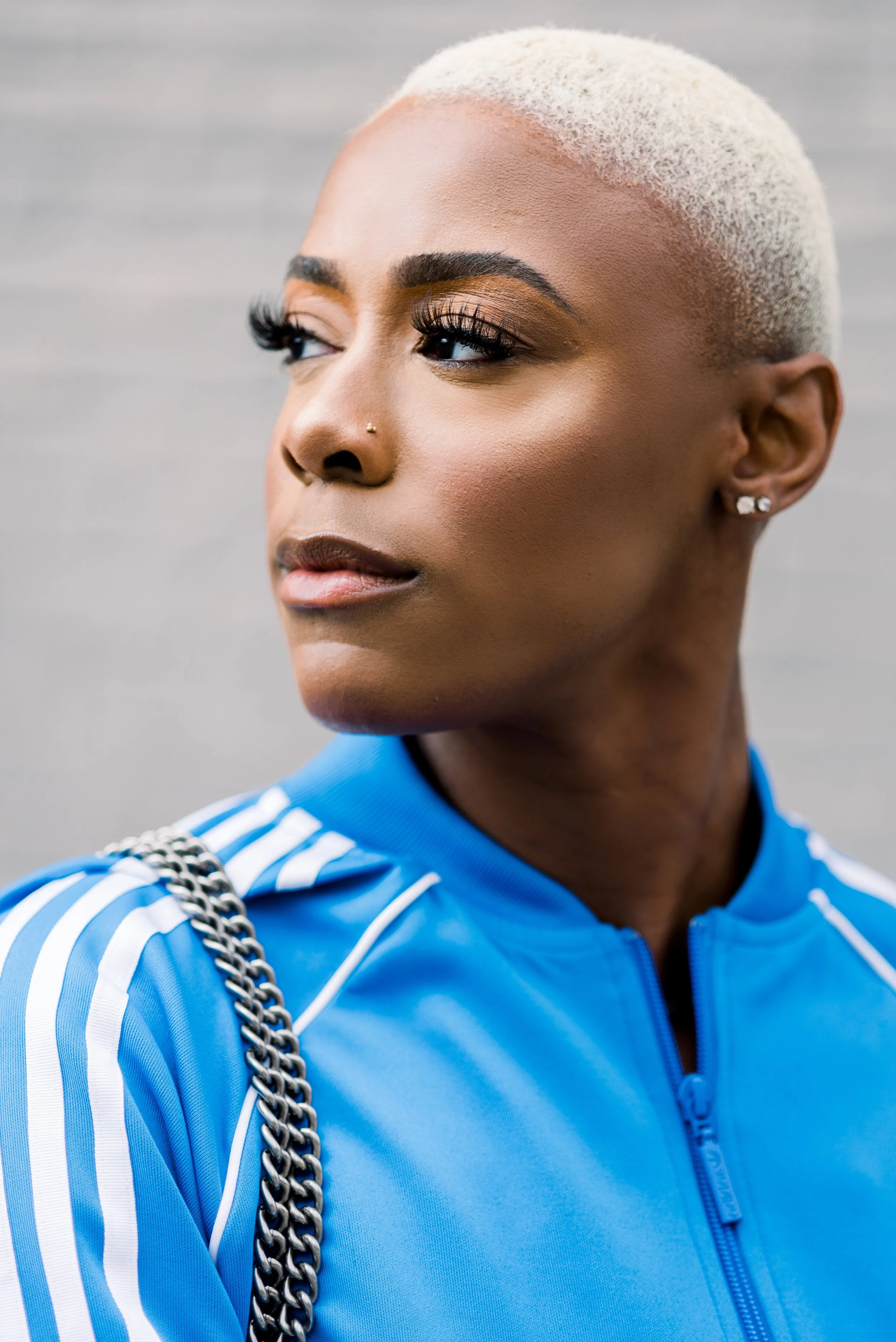 Close-up of a woman with short platinum blonde hair wearing a blue Adidas sports jacket, with a chain strap over her shoulder, standing against a gray background.