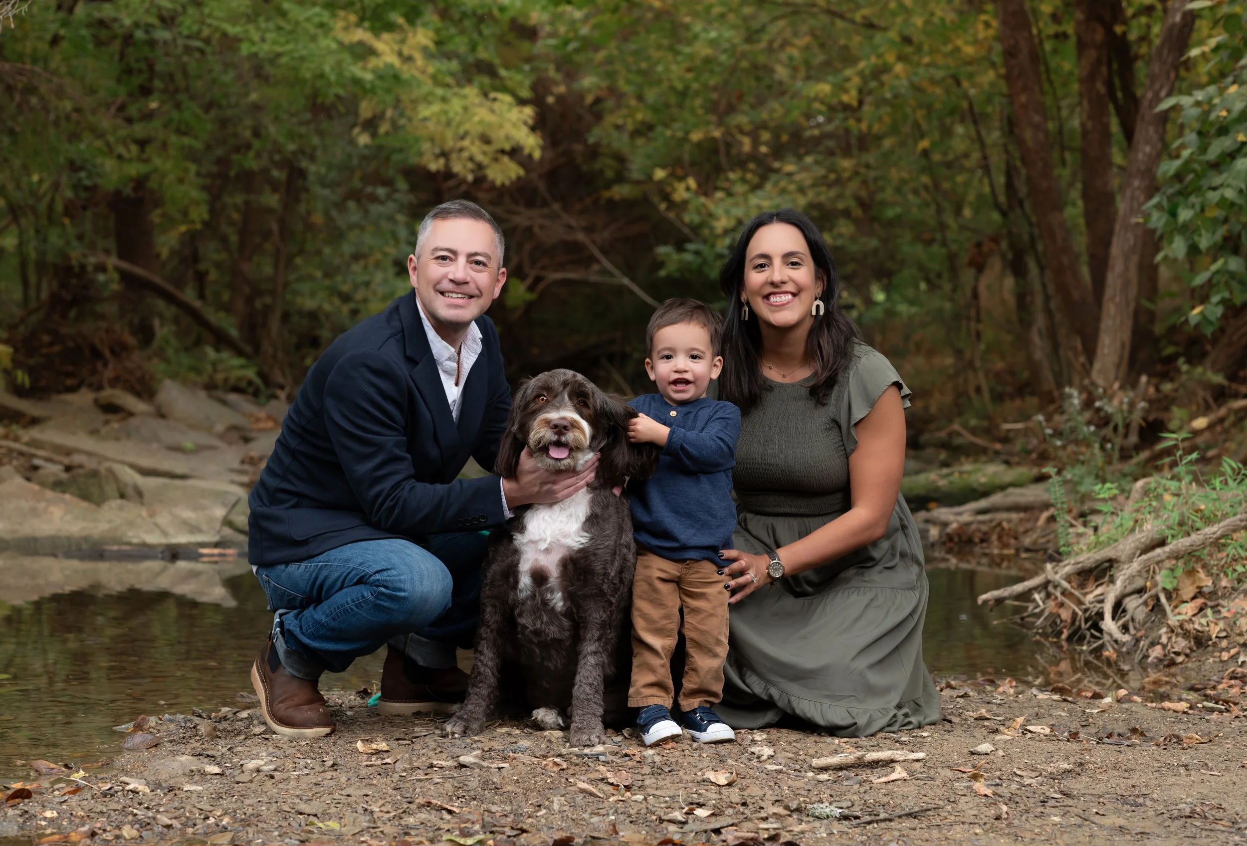 A family of four with a dog outdoors near a creek in a wooded area, smiling at the camera.